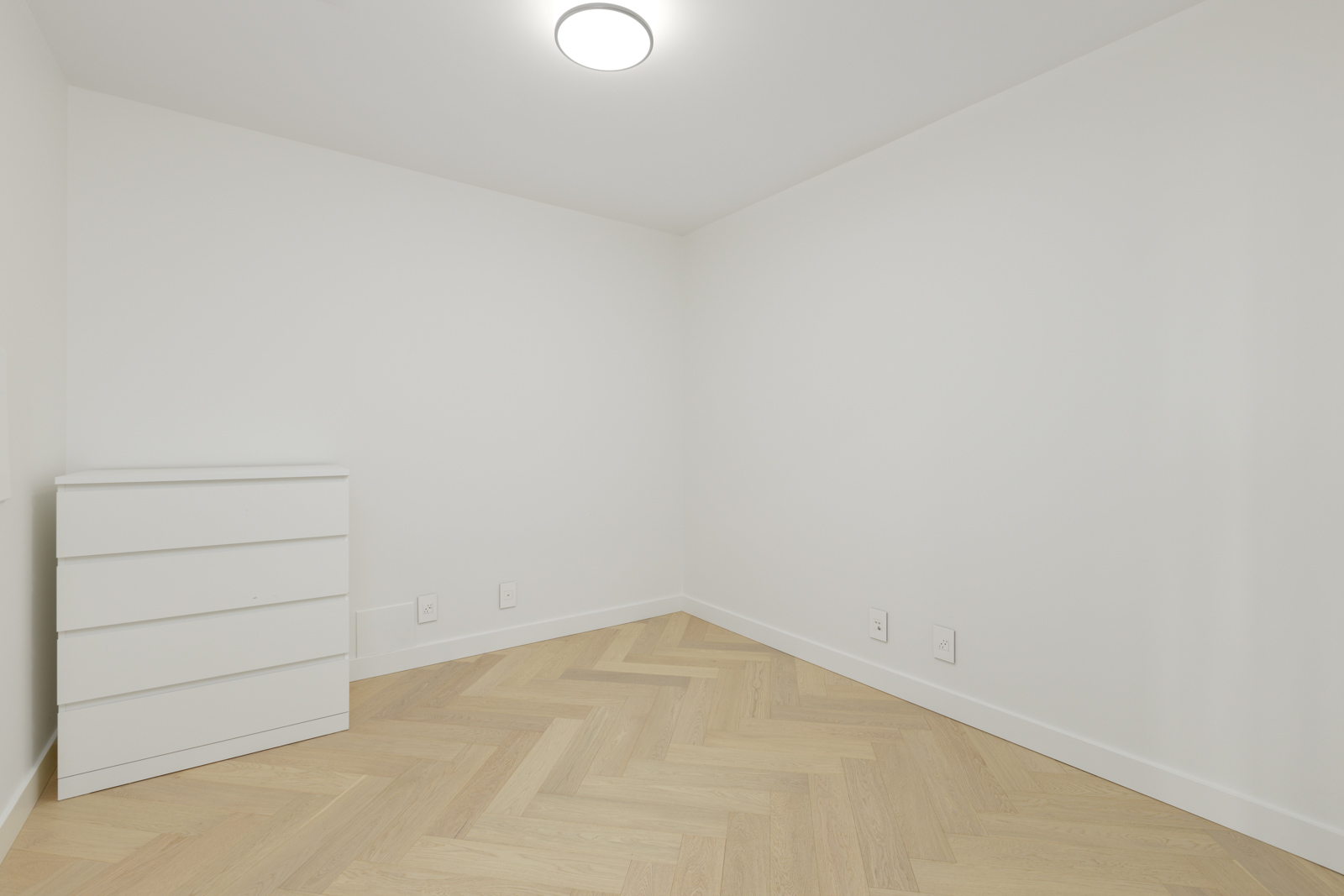 A small, empty room with white walls, light wood herringbone floor, a white dresser, and a round ceiling light.