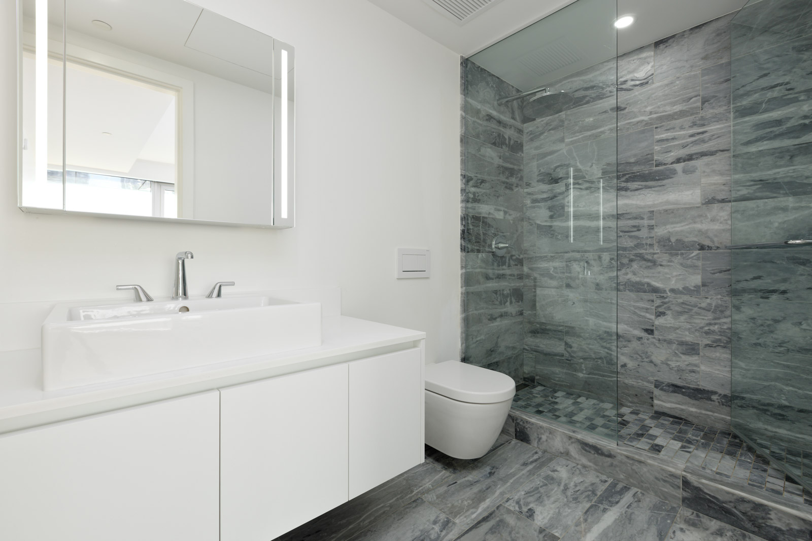 Modern bathroom with a white vanity, rectangular sink, wall-mounted toilet, and a glass-enclosed shower with gray marble walls and floor tiles.
