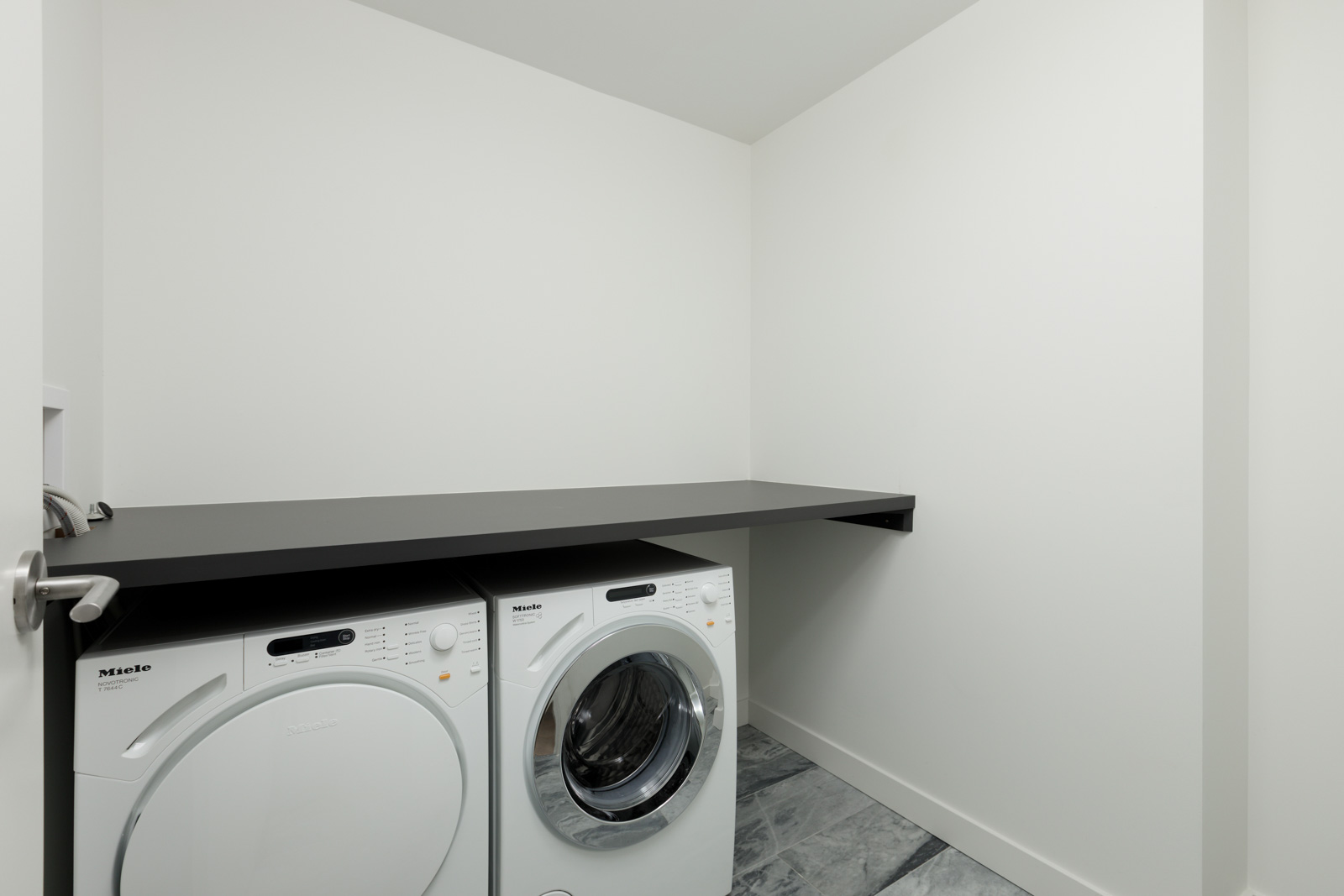 A modern laundry room with a white front-loading washer and dryer beneath a black countertop, set against plain white walls and a tiled floor.