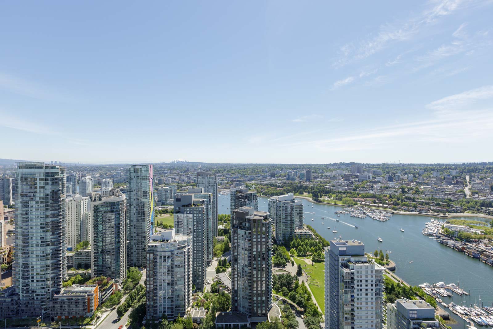 Aerial view of a cityscape with modern high-rise buildings, a green park, and a river with boats under a clear blue sky.