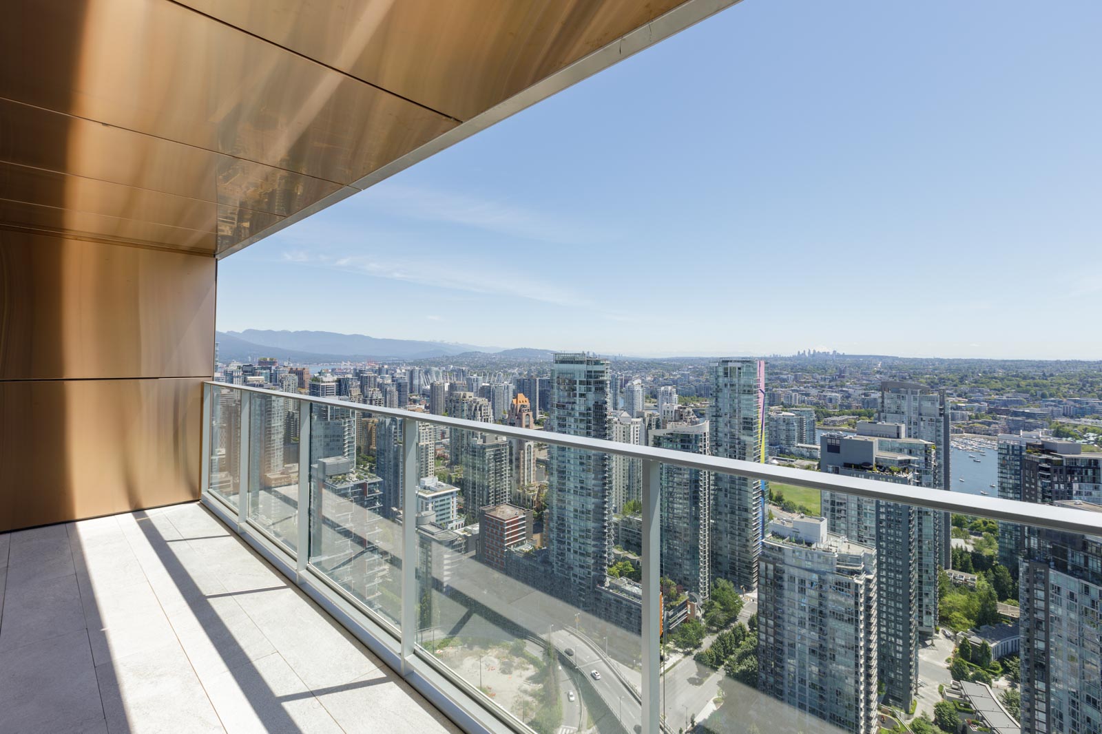 View from a high-rise balcony overlooking a cityscape with numerous tall buildings, glass railings, and distant mountains under a clear blue sky.