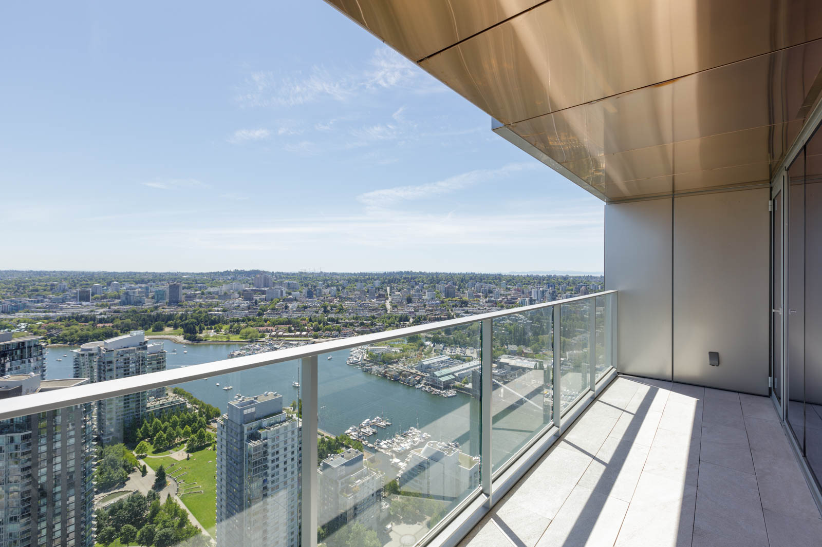 High-rise balcony with glass railing overlooking a cityscape, marina, and river under a clear blue sky.
