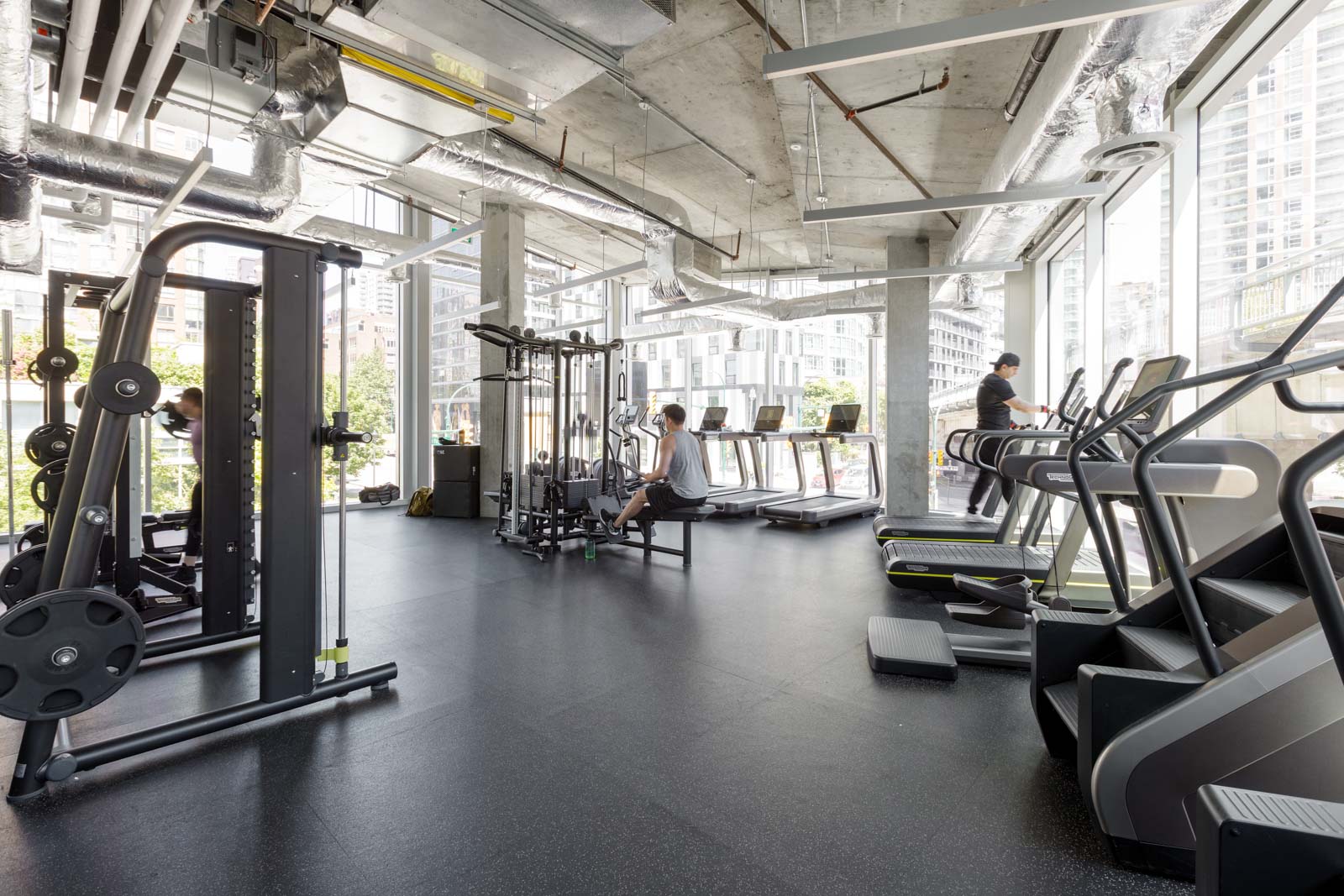 Modern gym with weight machines, treadmills, and exercise equipment. Two people are working out, one on a machine and another on a treadmill by the windows.