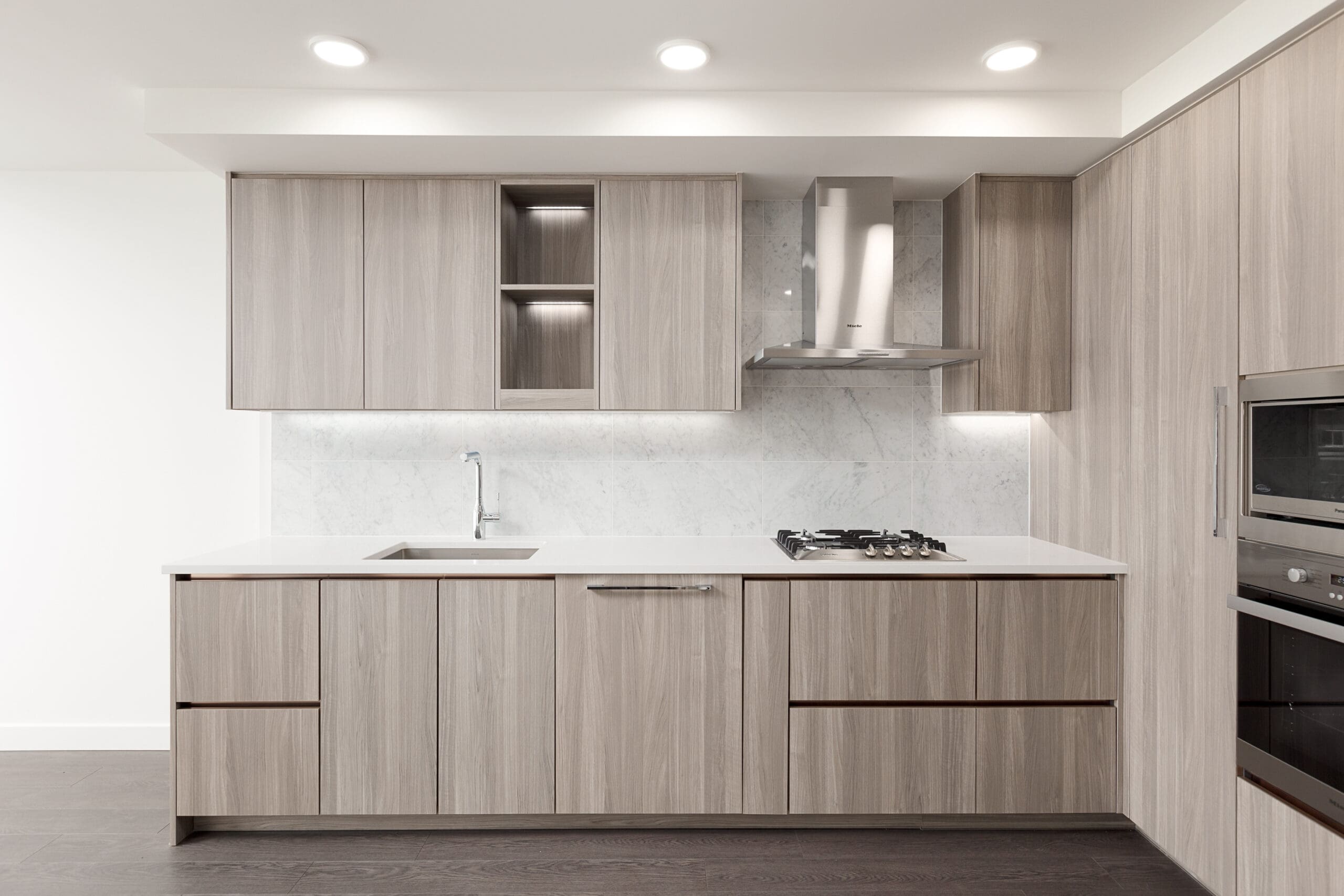 Modern kitchen with light wood cabinetry, built-in appliances, a gas cooktop, under-cabinet lighting, and a marble backsplash.