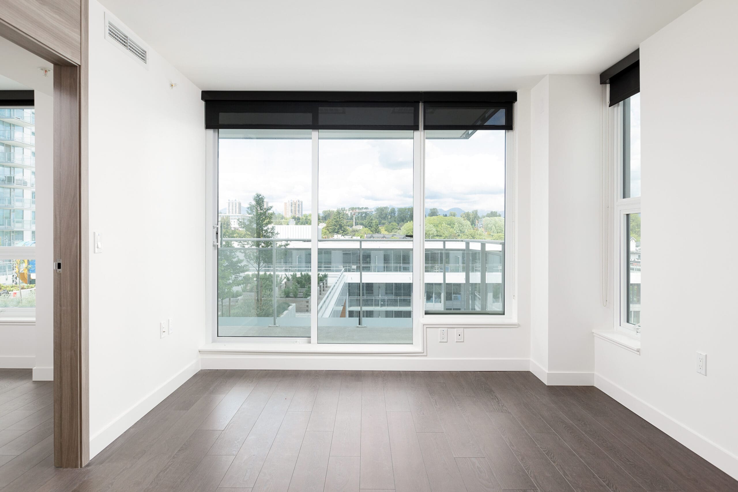 Bright, empty room with dark wood floors, large floor-to-ceiling windows, and a view of modern buildings and greenery outside.