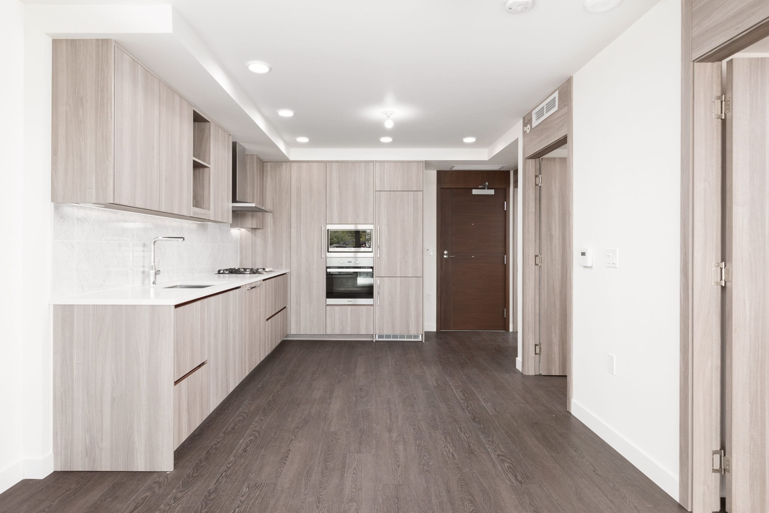 Modern, unfurnished kitchen with light wood cabinets, white countertops, built-in appliances, and dark wood floors, adjacent to an open entryway and closed doors.