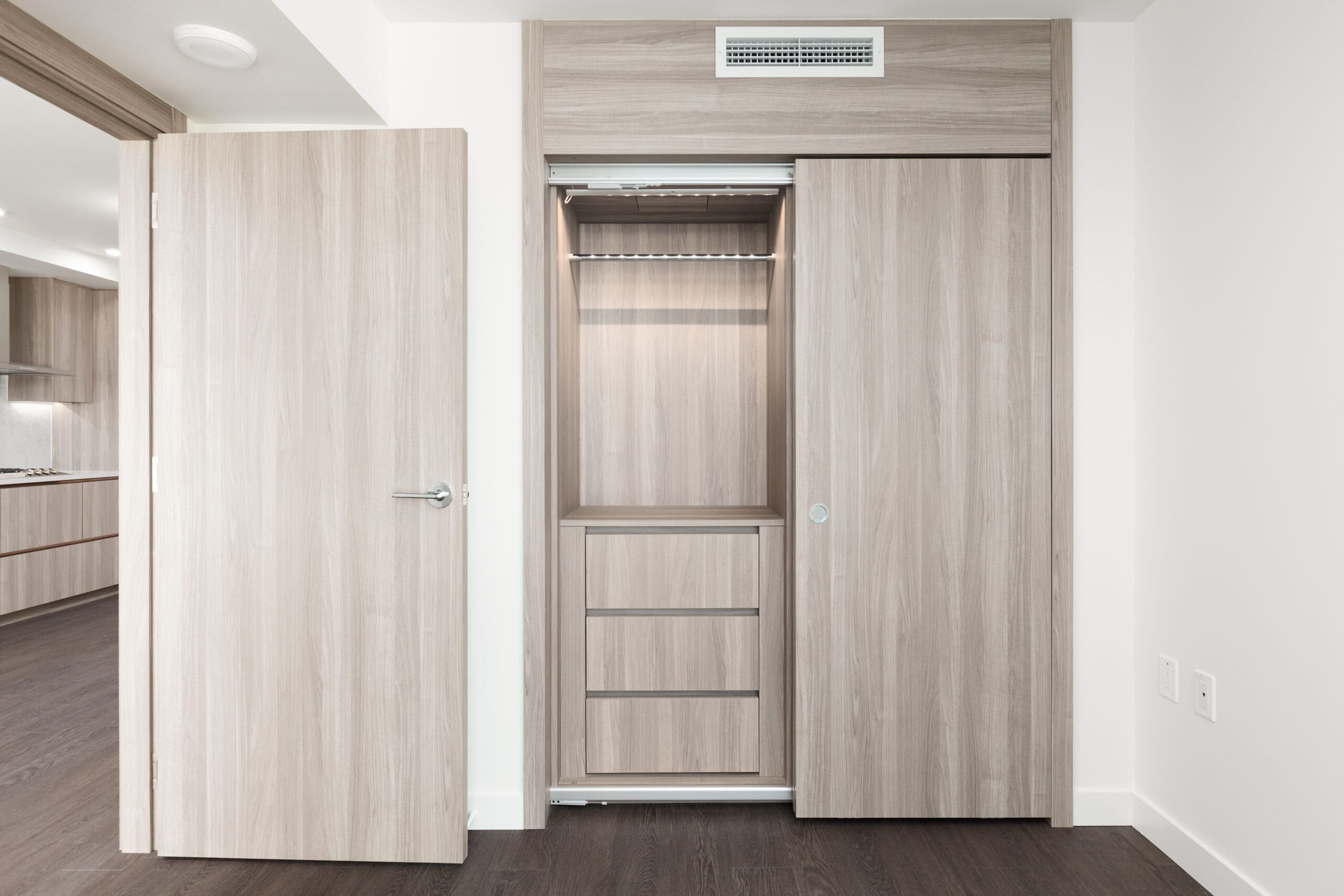 Open closet with sliding doors in a modern room, featuring hanging space, shelves, and three drawers. An adjacent hinged door opens to a view of a kitchen area.