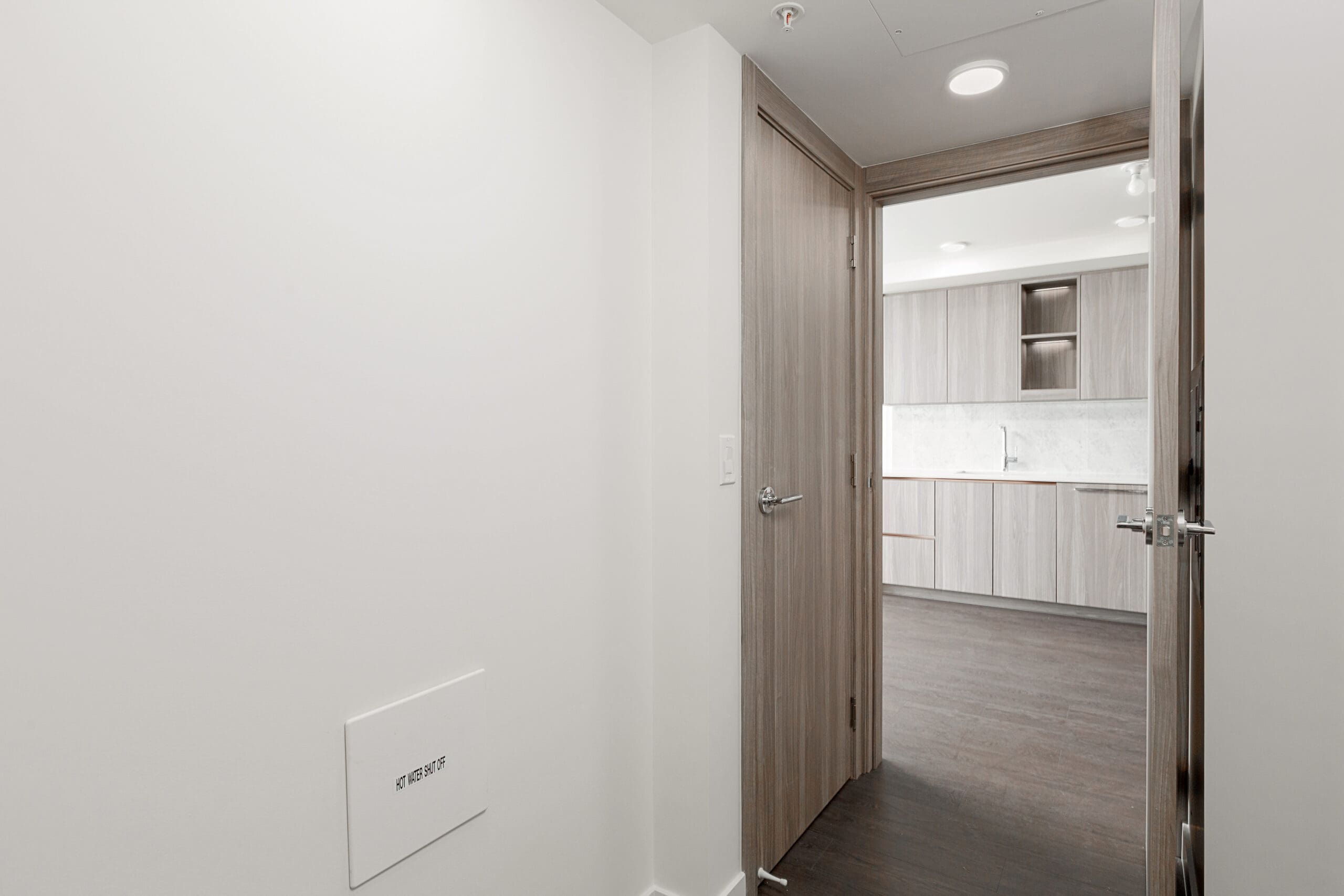 View of a hallway with light wood doors, leading into a modern kitchen with light wood cabinets, white countertops, and dark wood flooring.