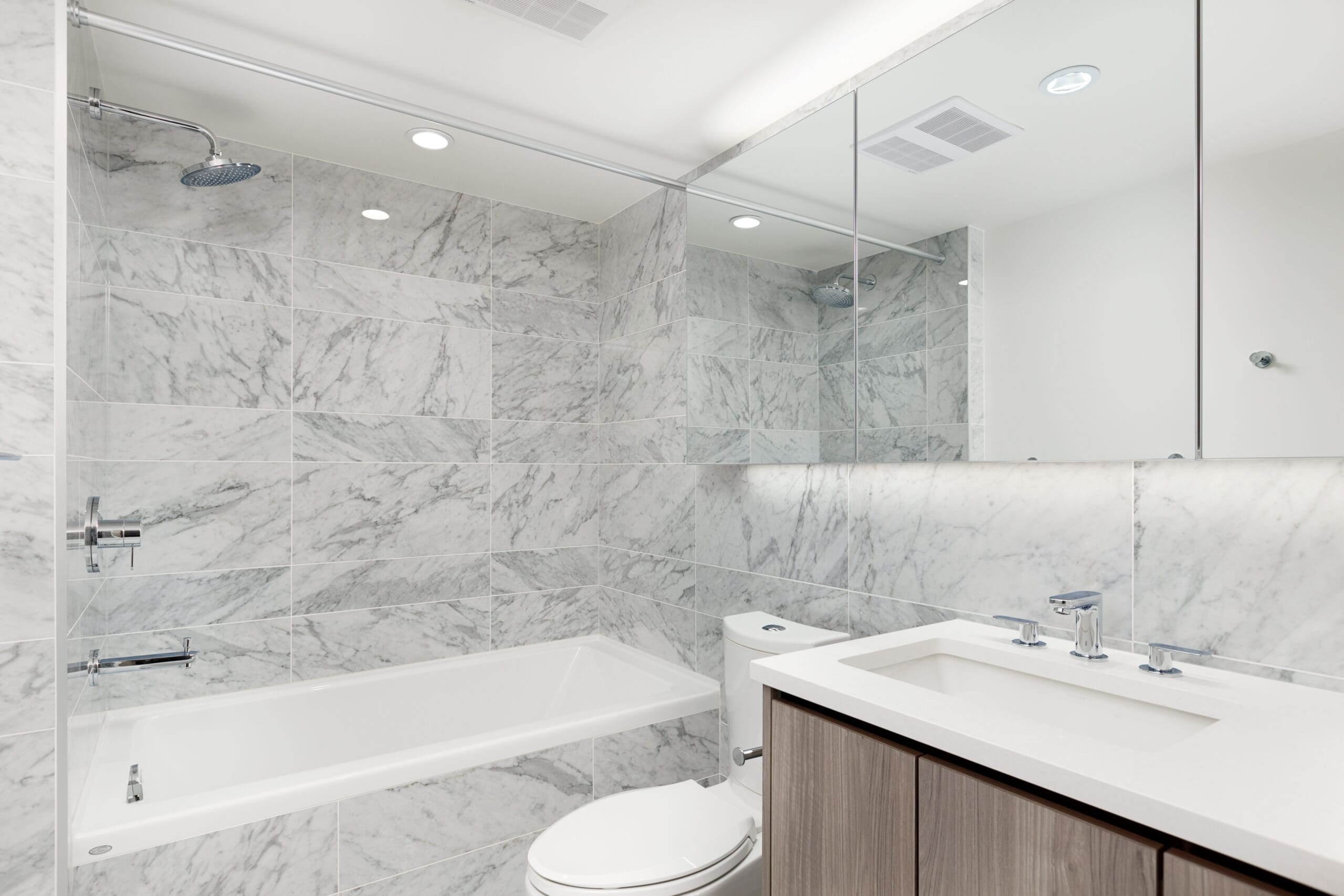 Modern bathroom with marble tile walls, a built-in bathtub, a wall-mounted showerhead, a toilet, a large mirror, and a double-sink vanity with a white countertop.