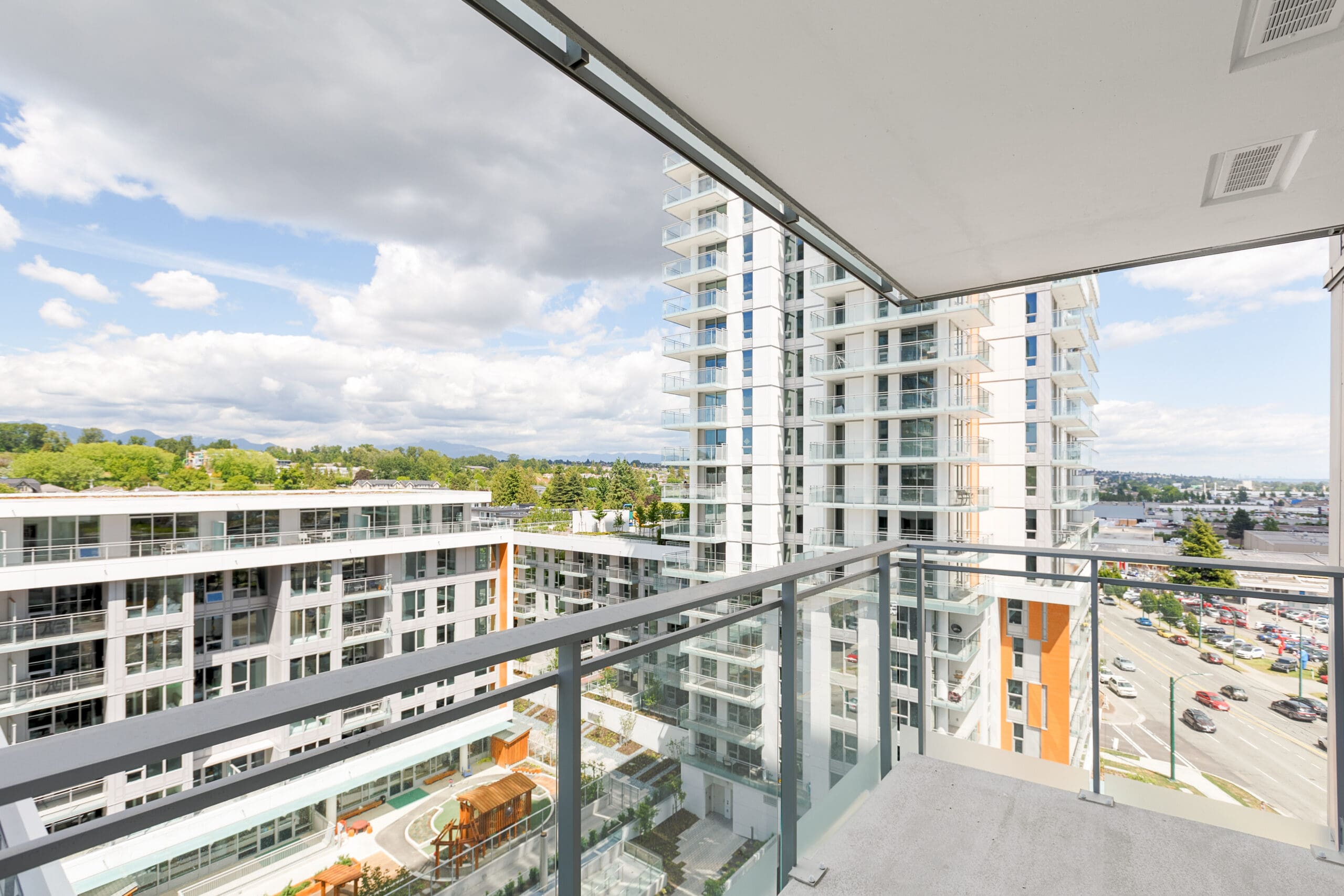 View from a modern apartment balcony overlooking other residential buildings, a parking lot, and a partly cloudy sky.