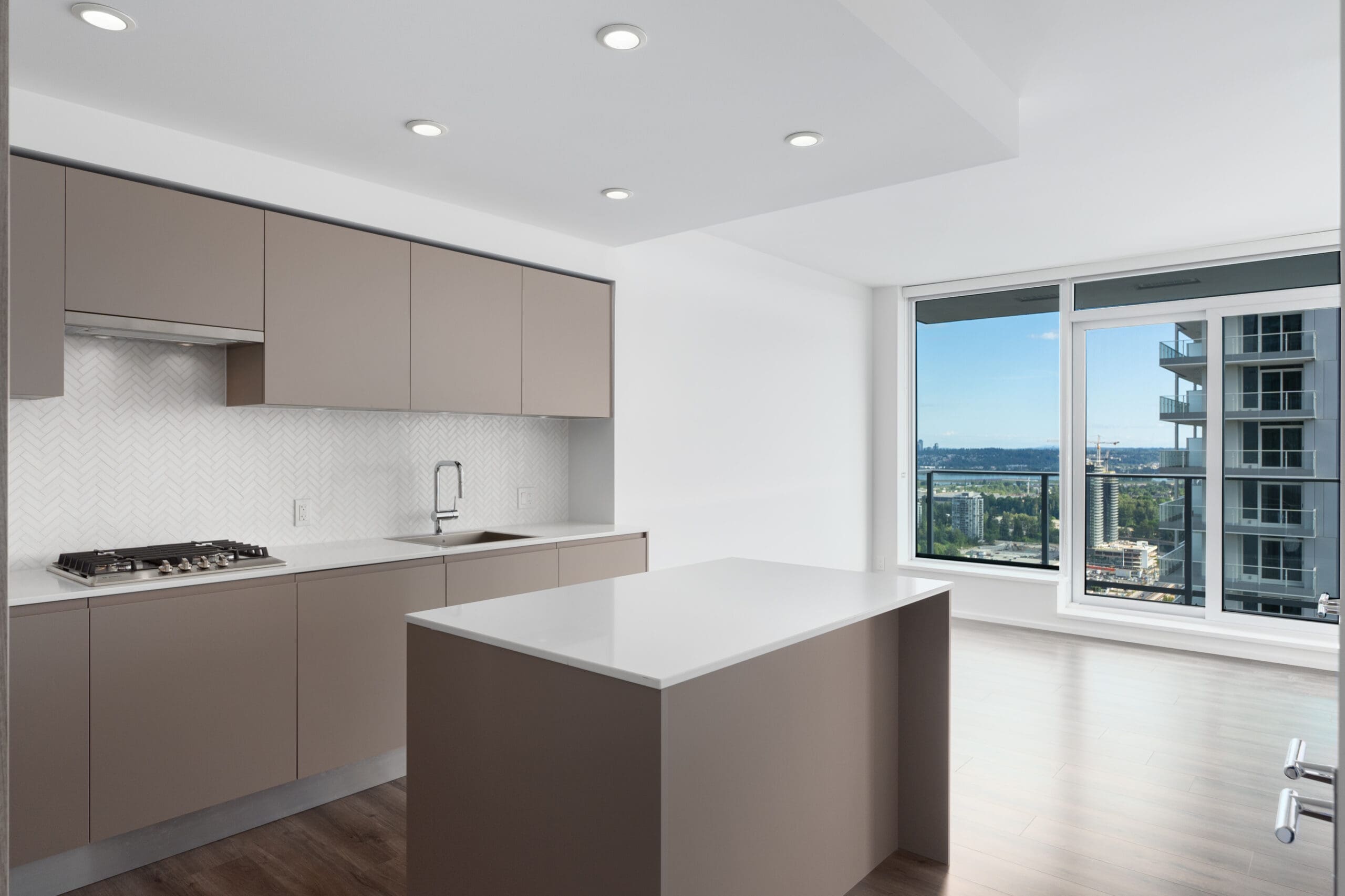 Modern kitchen with brown cabinets, white island, built-in stove, and large window overlooking cityscape and balcony.
