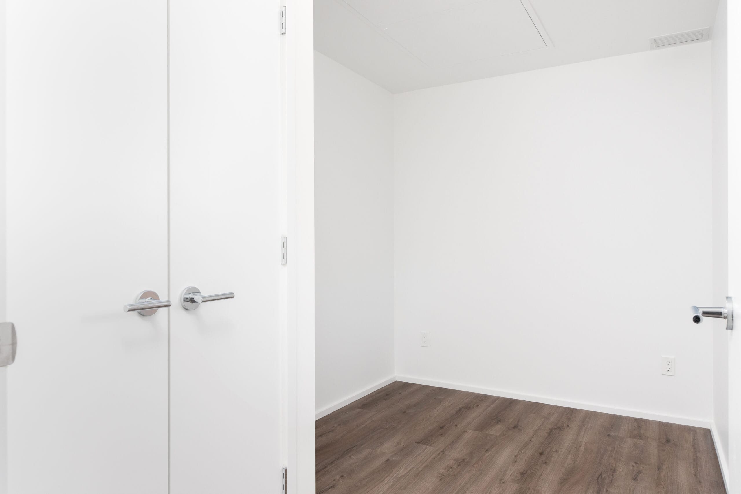 Empty, small white room with closed double doors, white walls, ceiling vent, electrical outlet, and dark wood flooring.