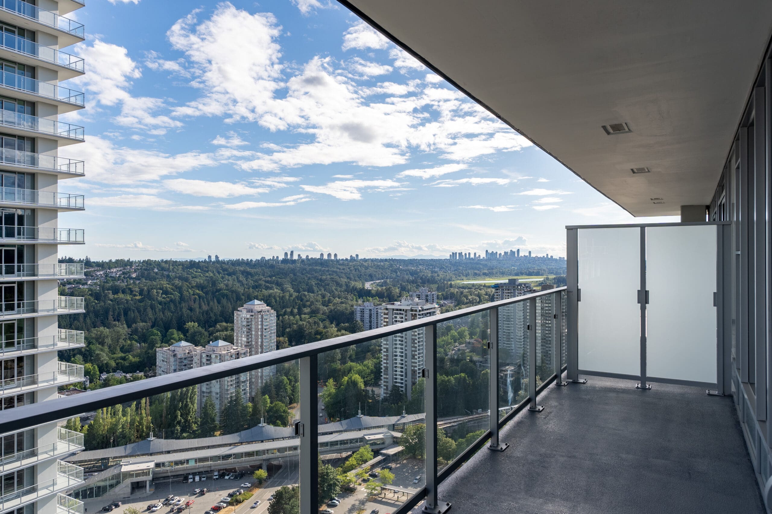 High-rise balcony with glass railing overlooking a cityscape, green forested area, and distant buildings under a partly cloudy sky.
