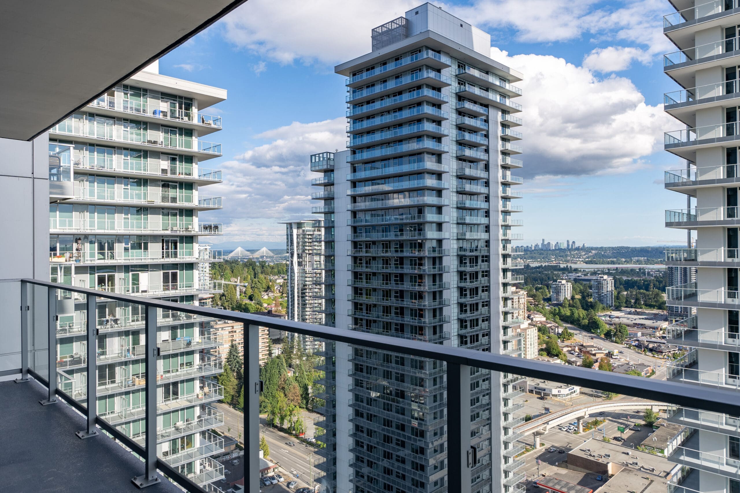 View from a high-rise balcony overlooking modern glass apartment buildings, city infrastructure, and a distant city skyline under a partly cloudy sky.