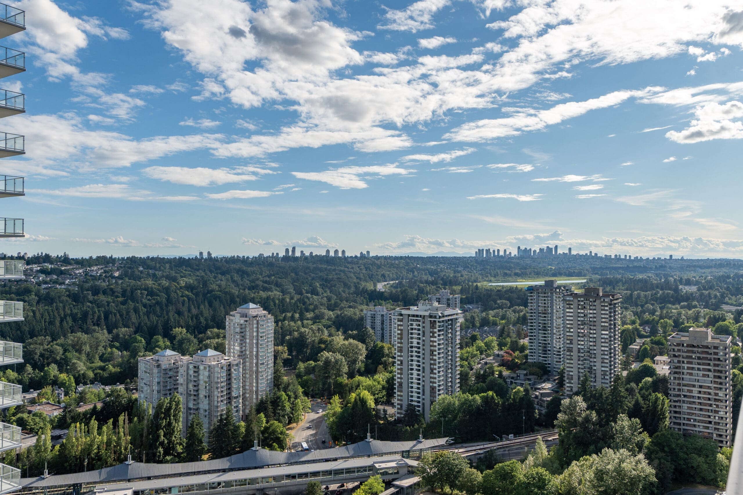 High-angle view of several high-rise apartment buildings surrounded by dense green trees, with a city skyline visible in the distant background under a partly cloudy sky.
