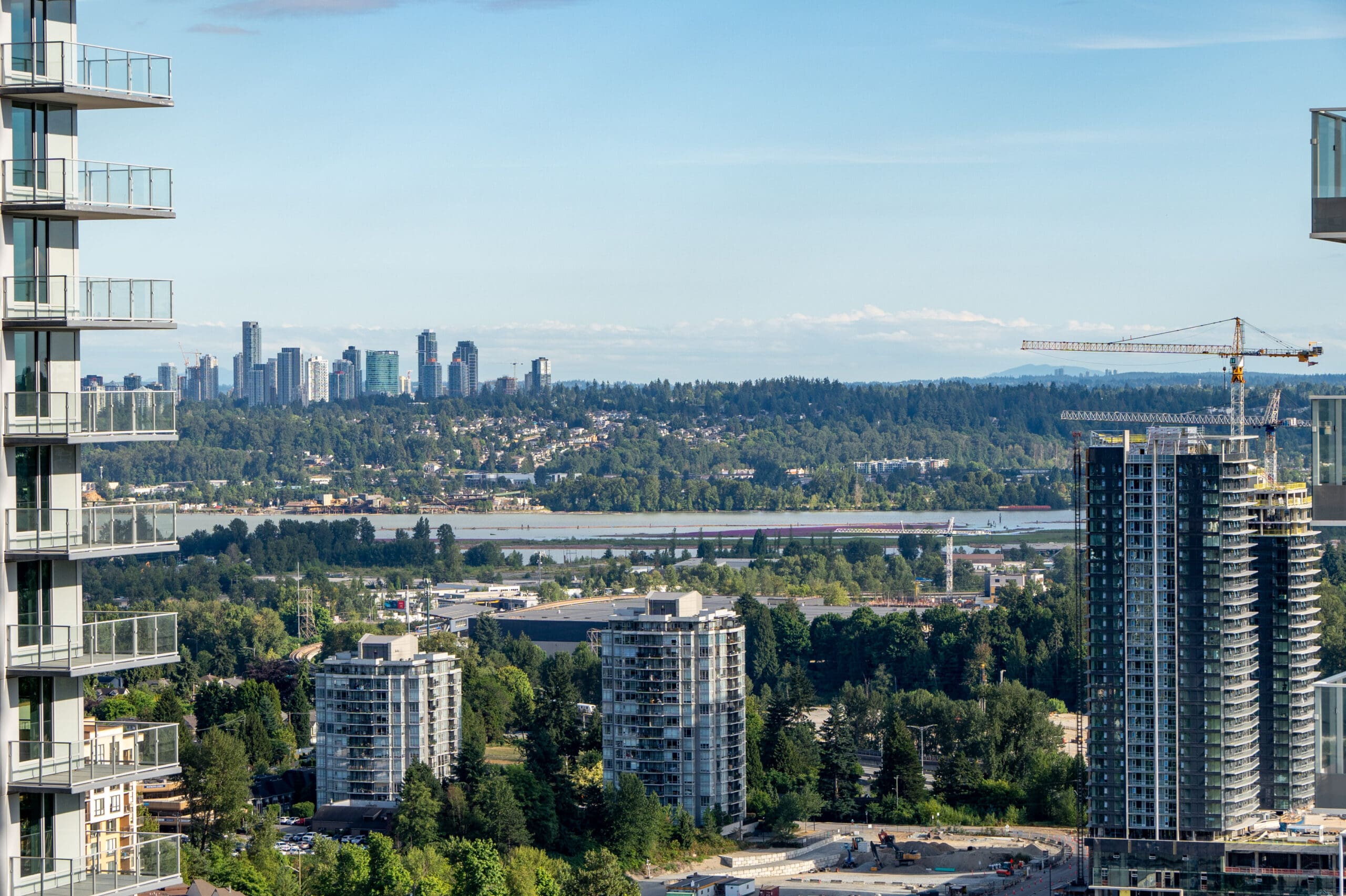 View of a cityscape with high-rise buildings, a construction crane, and a forested area in the distance under a clear blue sky.