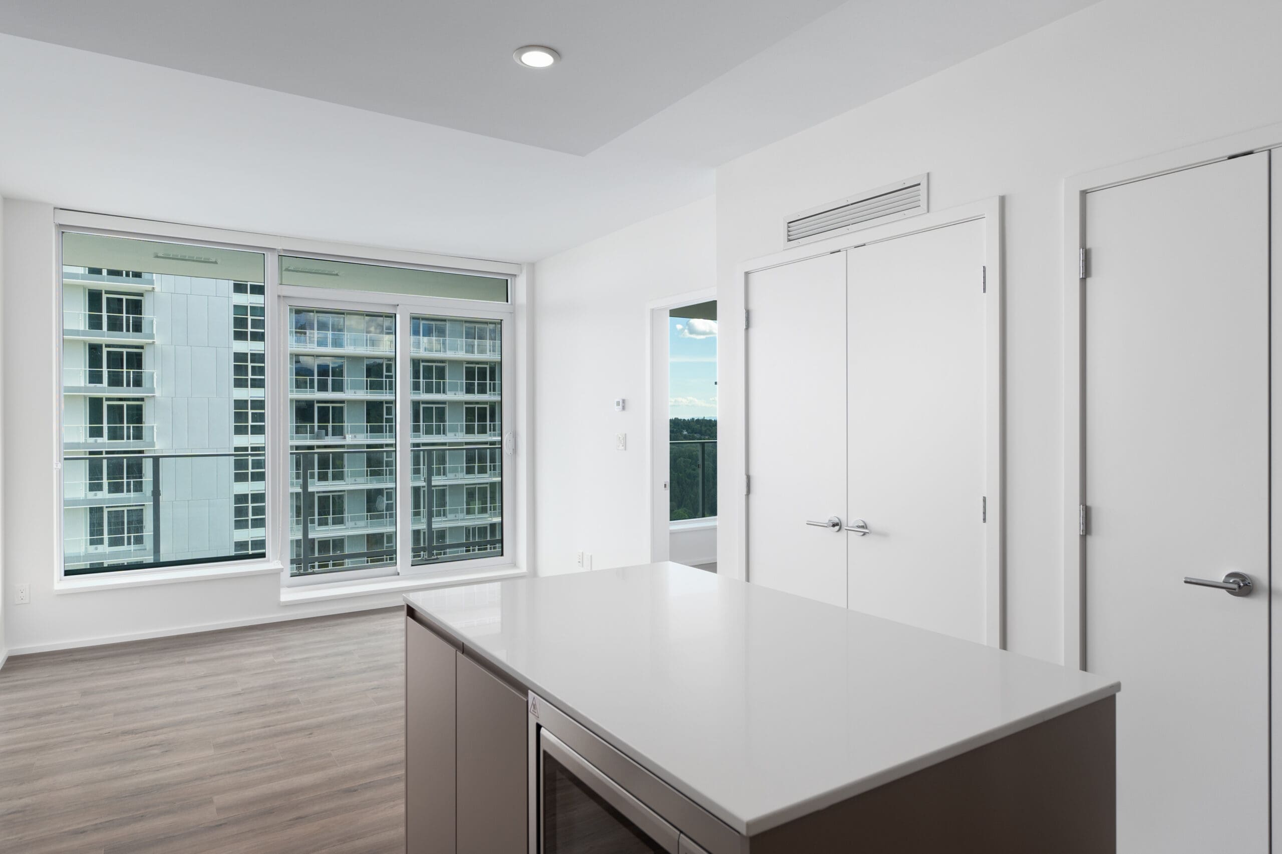 Modern apartment interior with a kitchen island, wood flooring, large windows, and white walls, overlooking a neighboring building and cityscape.
