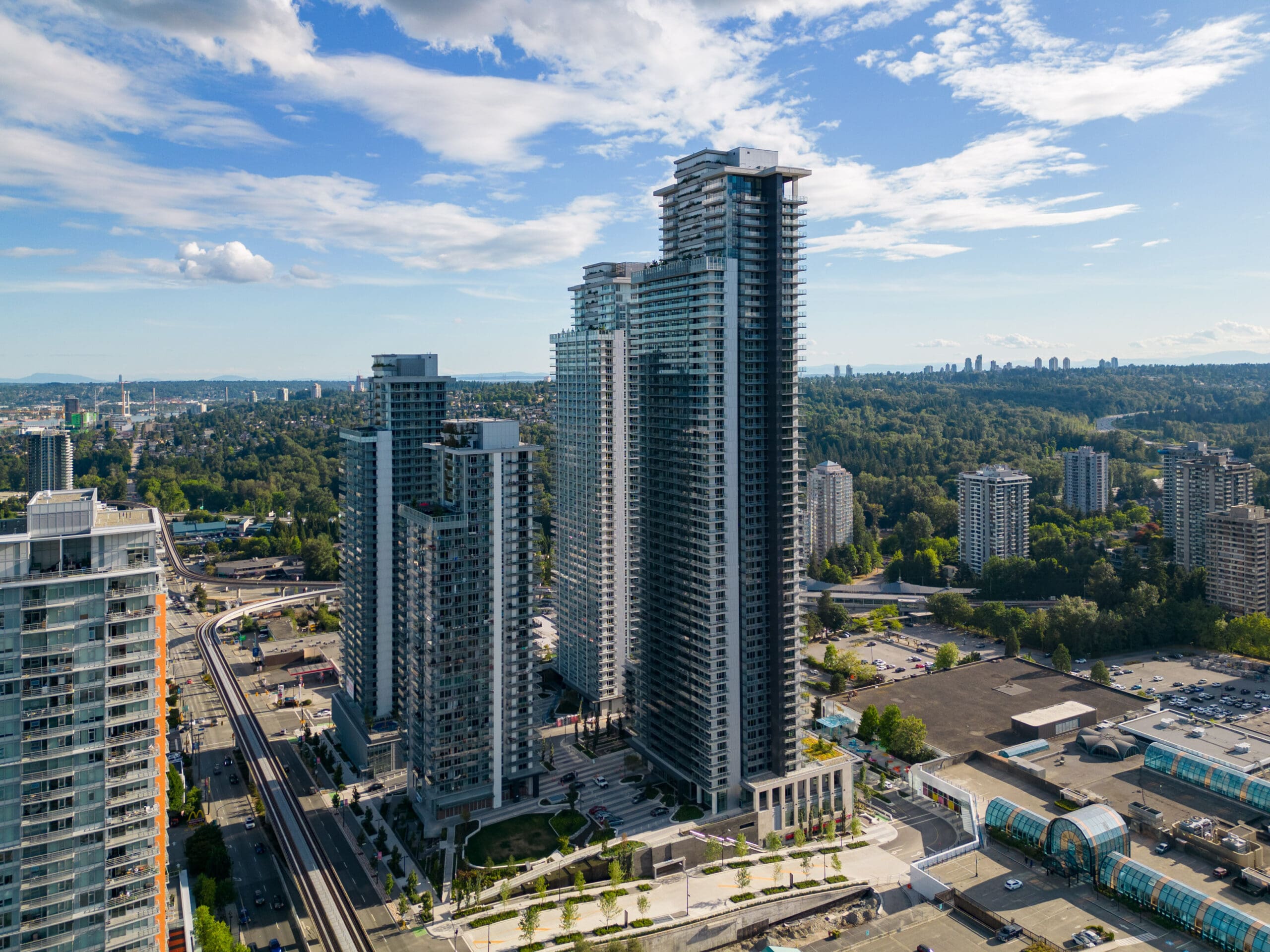 Aerial view of several tall modern condominium buildings surrounded by trees, roads, and commercial structures under a partly cloudy sky.