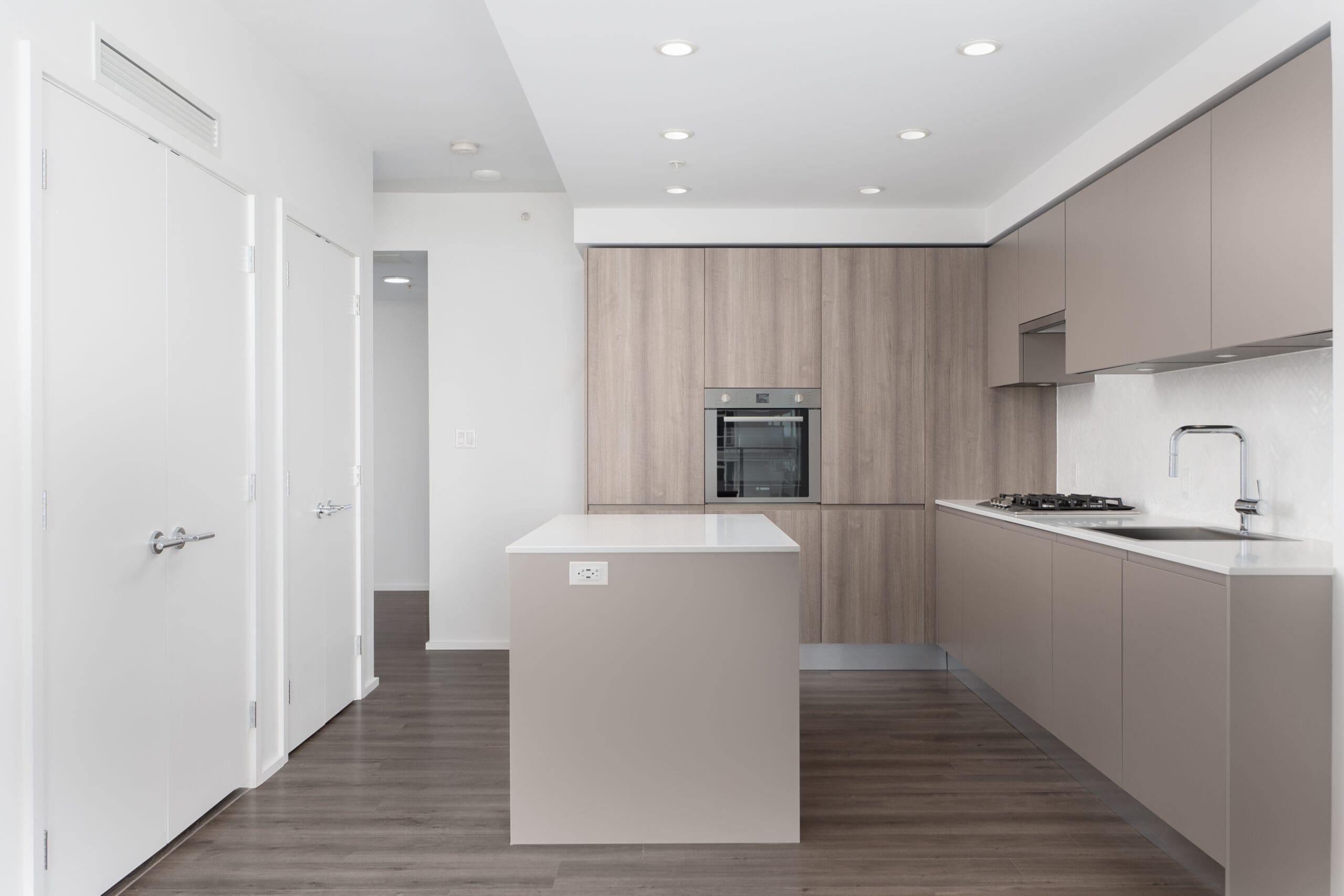 Modern kitchen with beige cabinets, built-in oven, gas cooktop, island with white countertop, and wood-look flooring. Walls and doors are white, ceiling has recessed lighting.