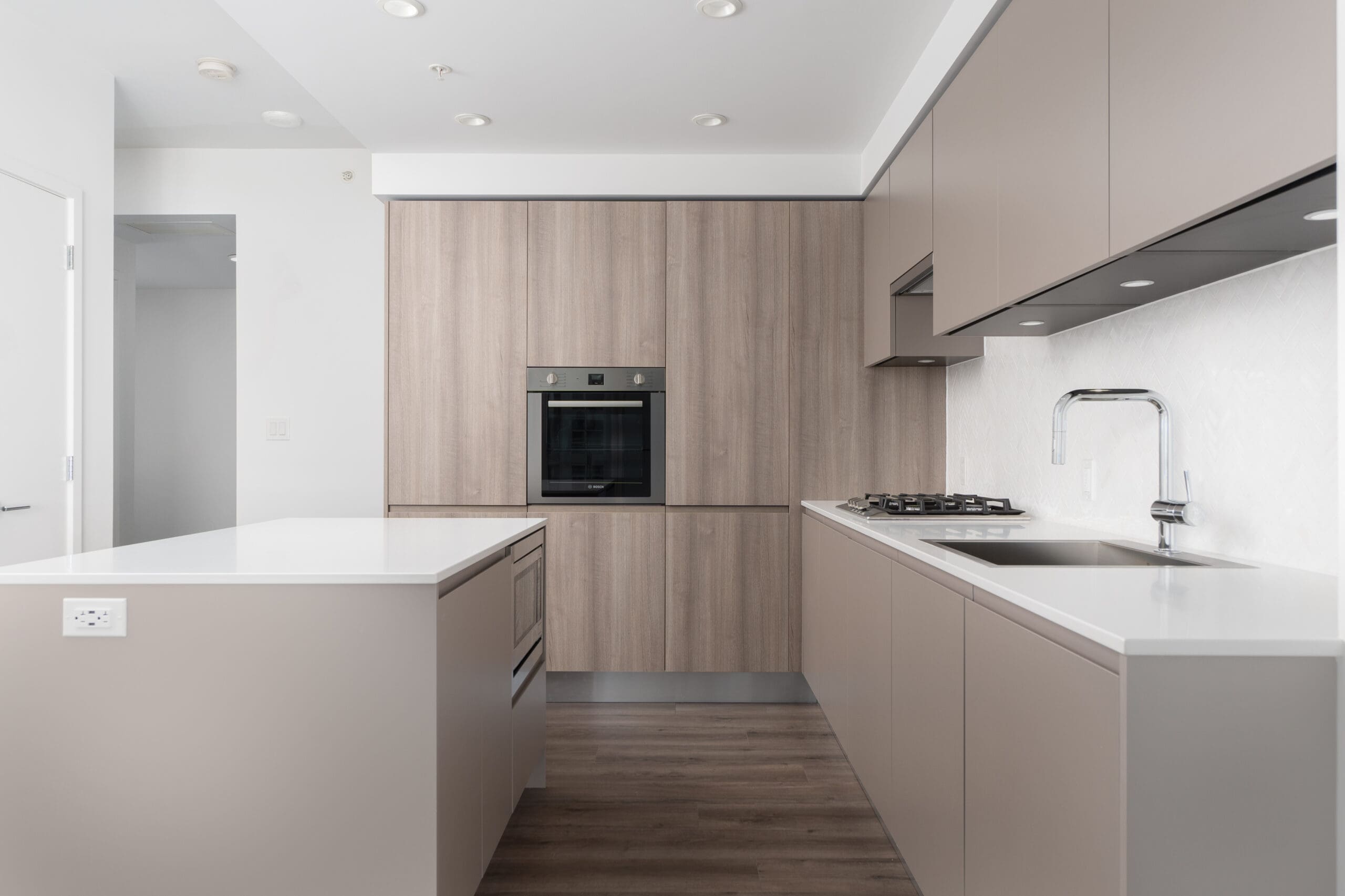 Modern kitchen with beige cabinets, built-in oven, gas cooktop, integrated sink, white countertops, and wood flooring.