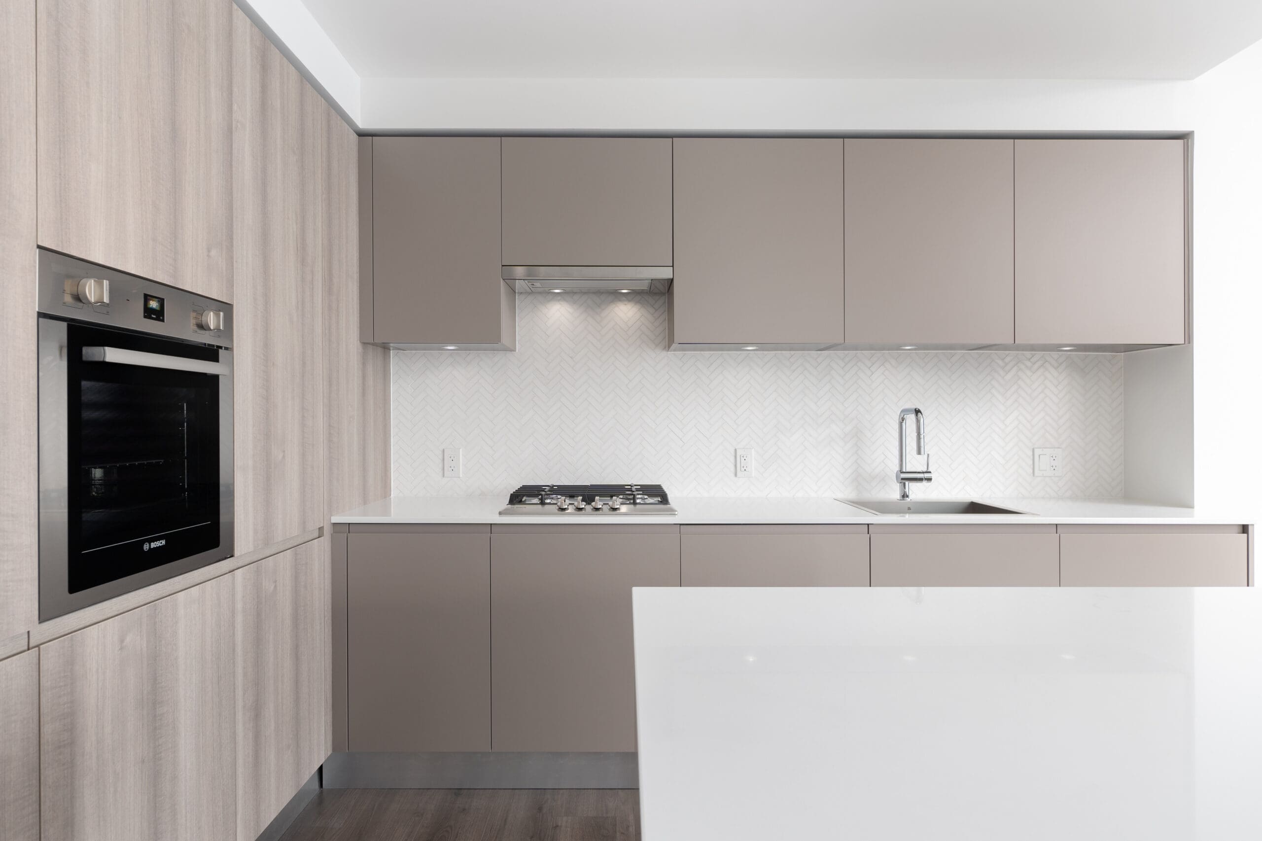 Modern kitchen with built-in oven, gas cooktop, sink, light wood and beige cabinets, white countertop, and herringbone tile backsplash.