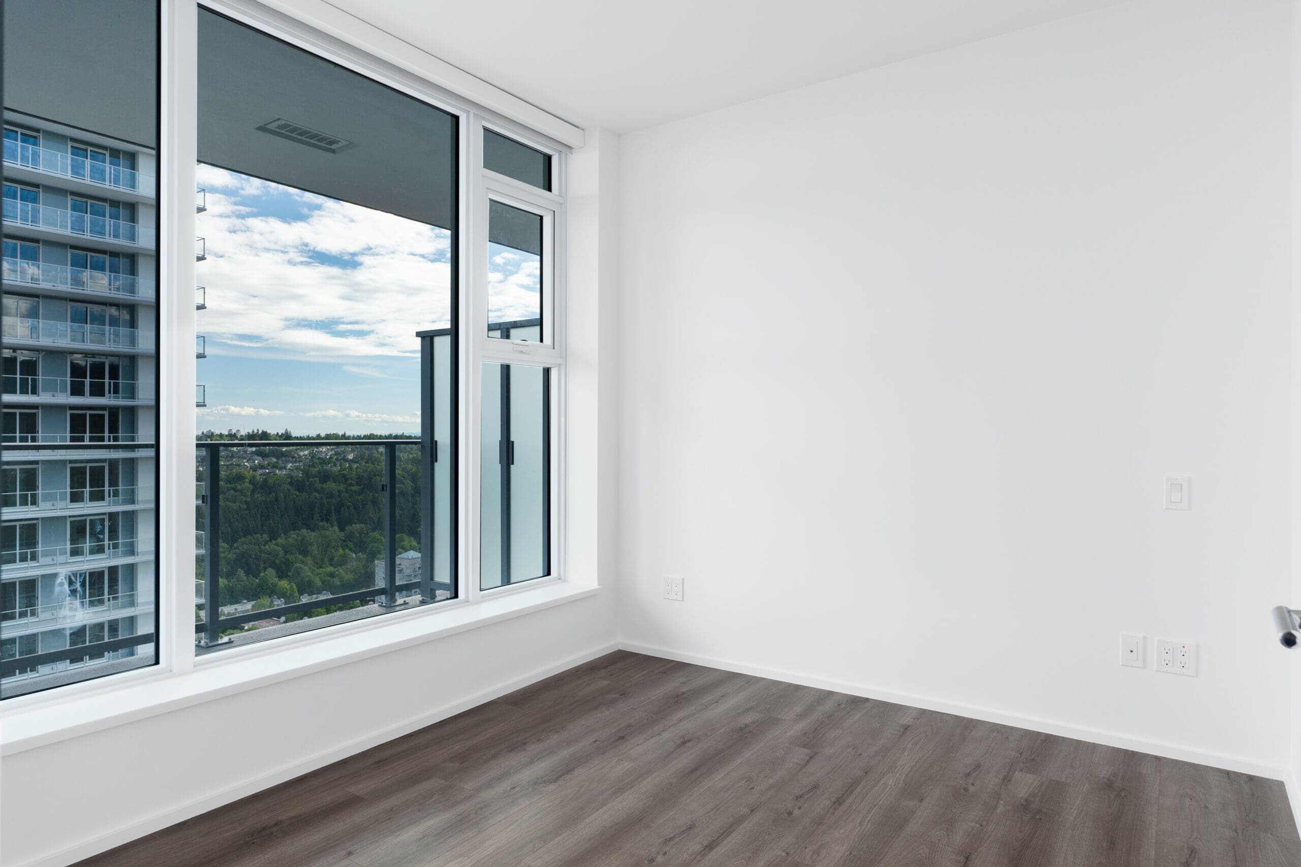 Empty room with white walls, large windows, and dark wood flooring. The windows offer a view of trees, a neighboring building, and a partly cloudy sky.