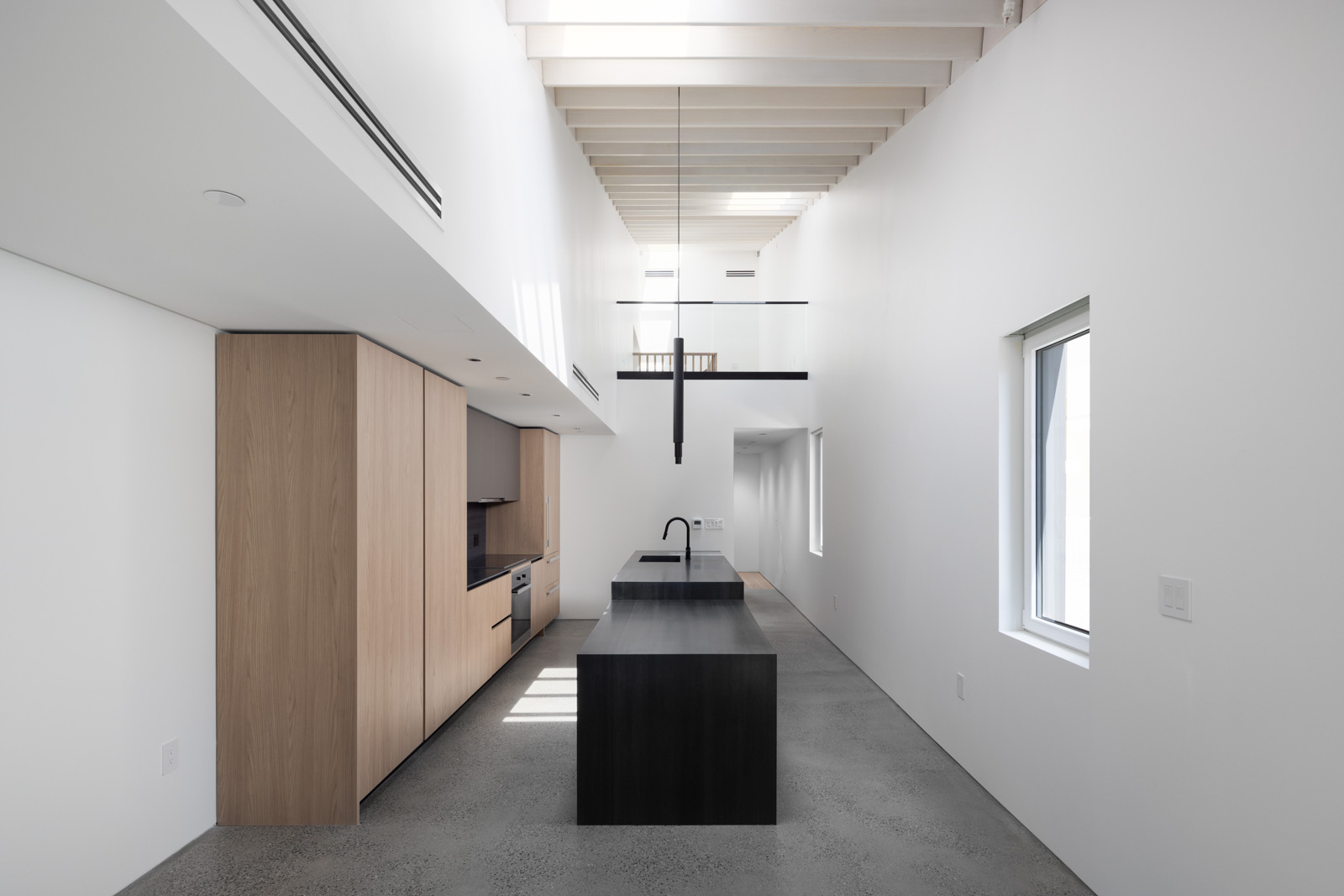 Minimalist modern kitchen with light wood cabinets, black countertop island, concrete floor, white walls, exposed ceiling beams, skylights, and a window.