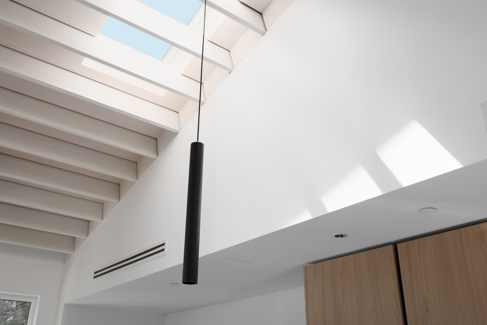 A black cylindrical pendant light hangs from a high ceiling with exposed beams and a skylight, letting sunlight into a modern, minimal interior.