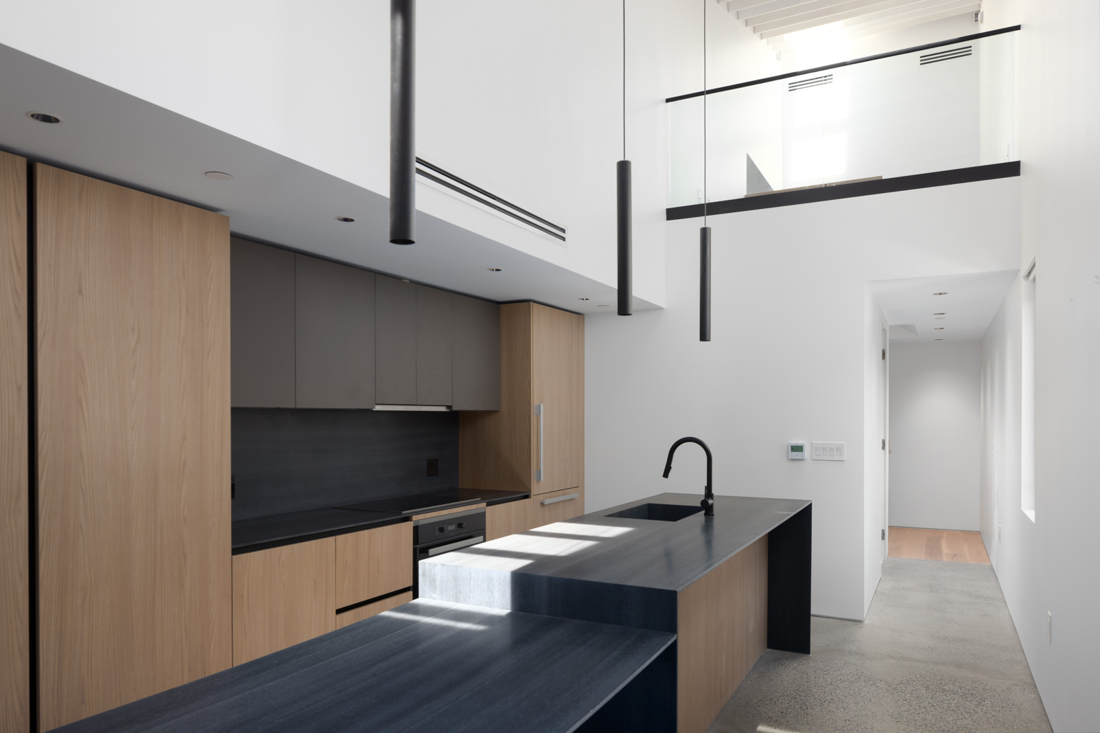 Modern kitchen with light wood cabinets, matte black countertops, built-in appliances, overhead pendant lights, and a hallway with glass railing on an upper level.