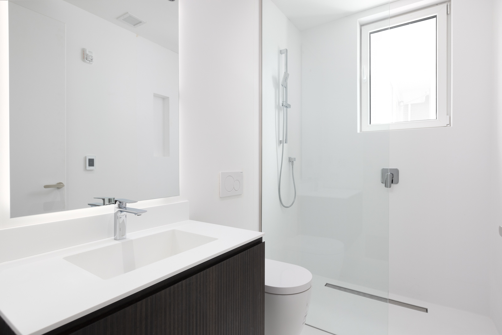 Minimalist bathroom with a white sink, dark vanity, wall-mounted faucet, glass shower enclosure, and a window letting in natural light.