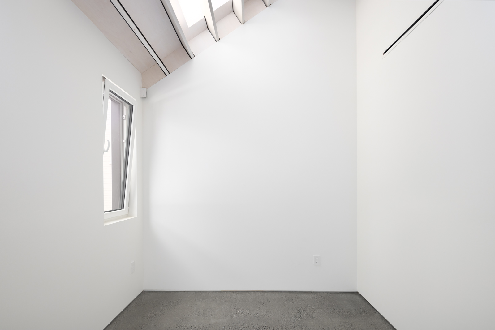 A small, empty white room with a single window on the left wall and a slanted ceiling with skylights letting in natural light.