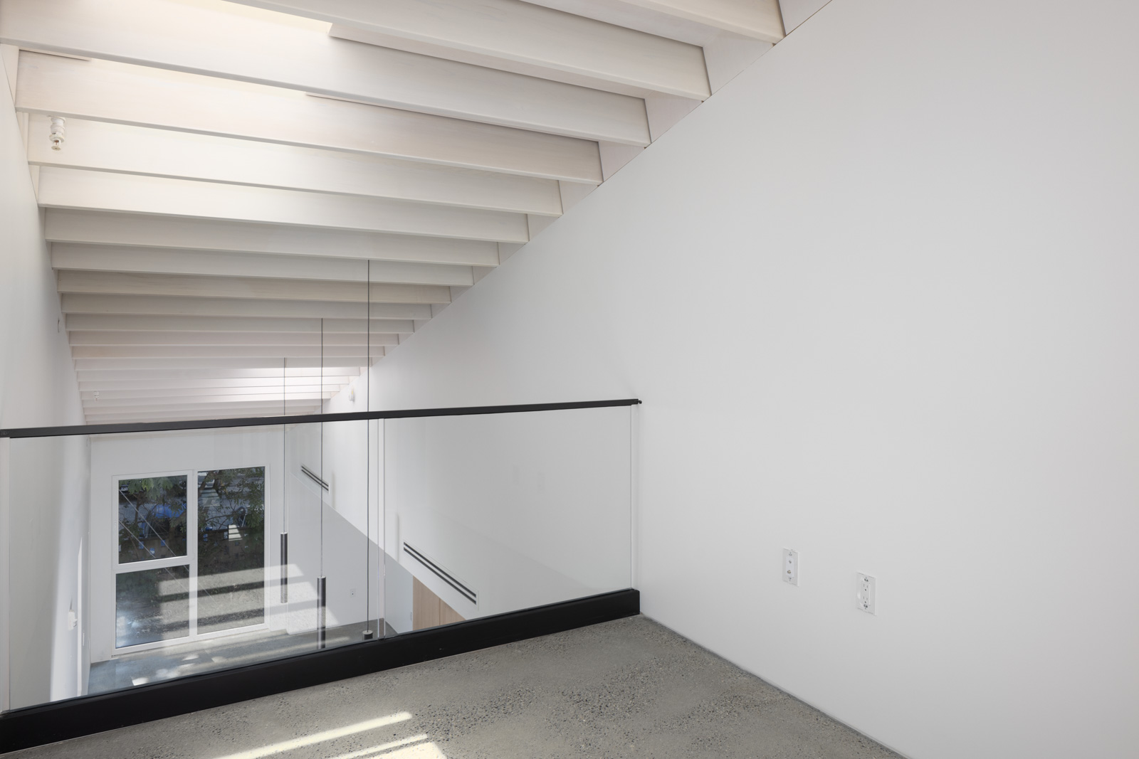 A modern loft area with exposed beams, glass railing, white walls, and a view of large windows overlooking an outdoor space.