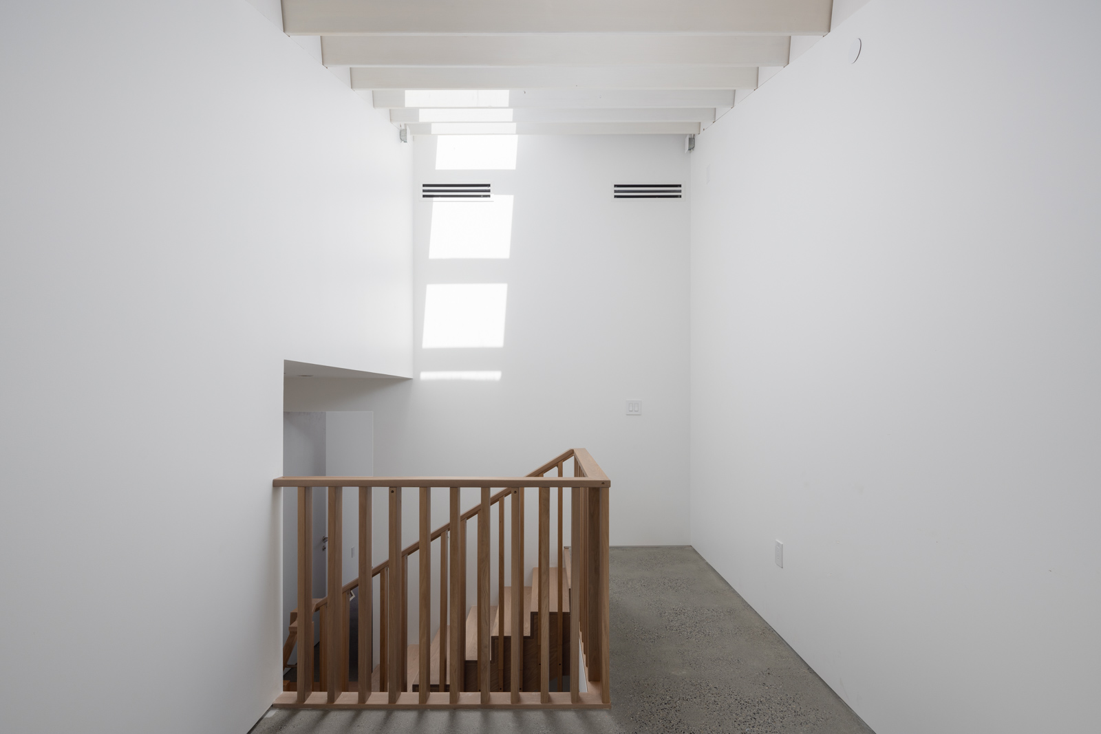 A wooden staircase with a simple railing leads down from a landing in a white, minimalist interior with exposed ceiling beams and a window casting rectangular light patterns on the wall.