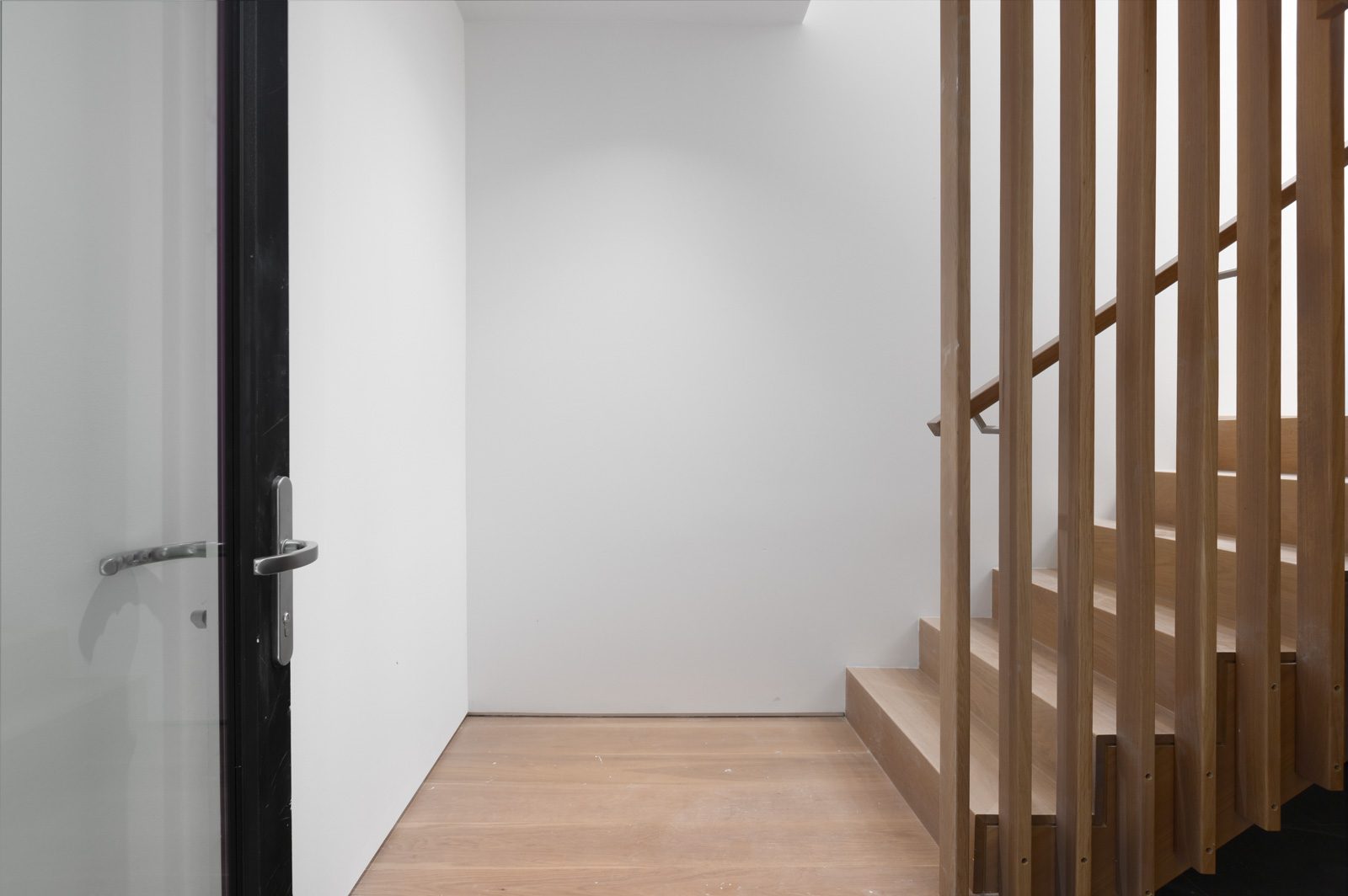 Minimalist interior with a wooden staircase on the right, vertical wooden slats, a glass door on the left, and white walls with a wooden floor.