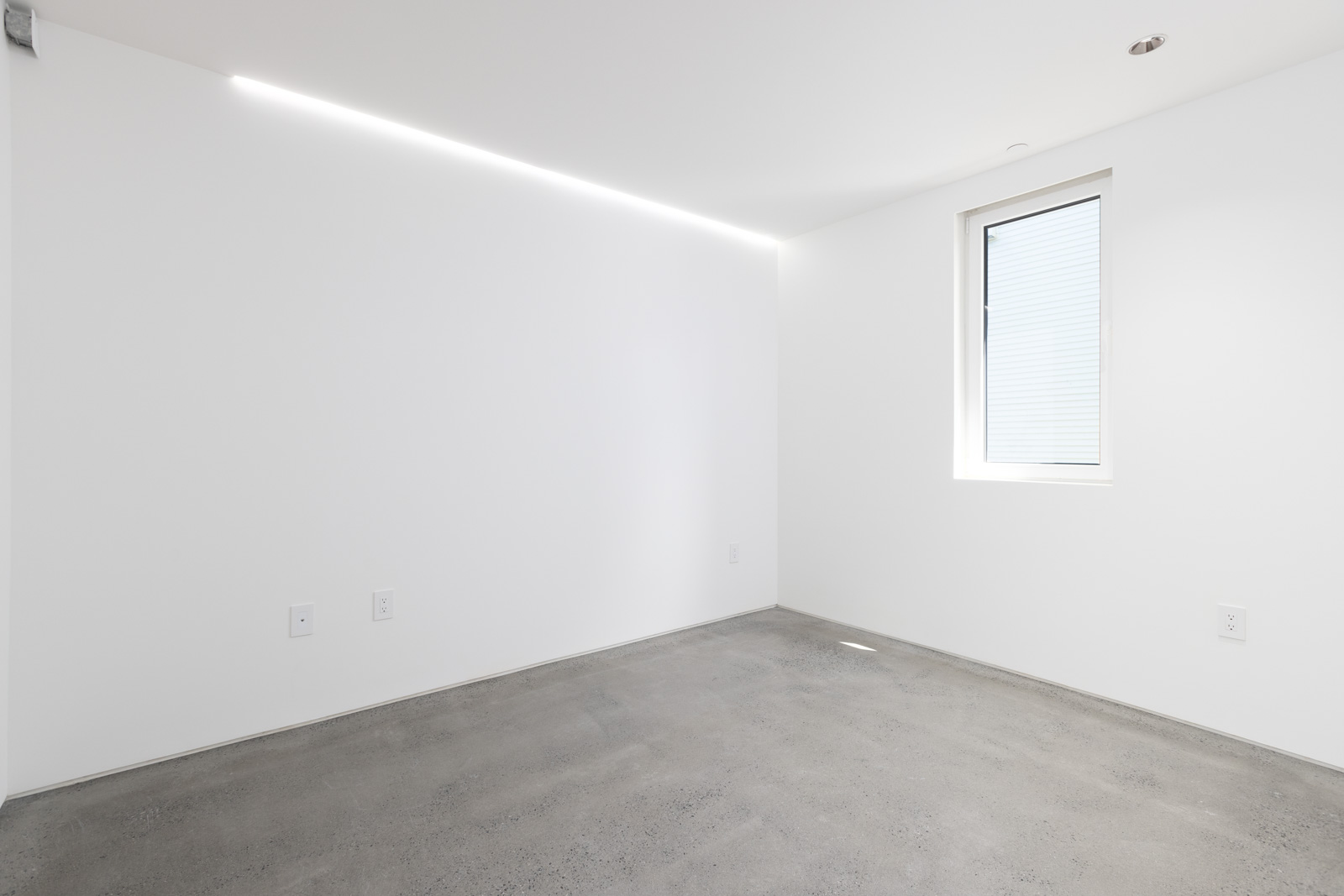 Minimalist empty room with white walls, a single window, multiple wall outlets, and a polished concrete floor. Ceiling light strip provides illumination.