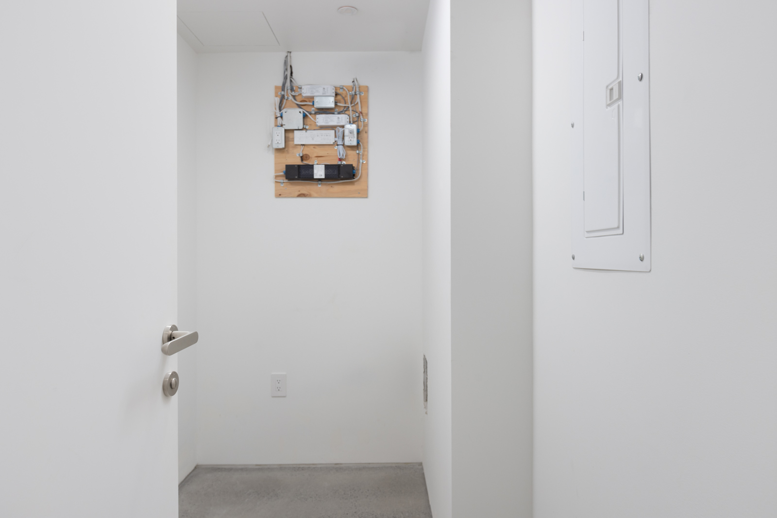 A small, empty white room with an open door, exposed wiring on a wooden board mounted on the wall, and an electrical panel on the right.