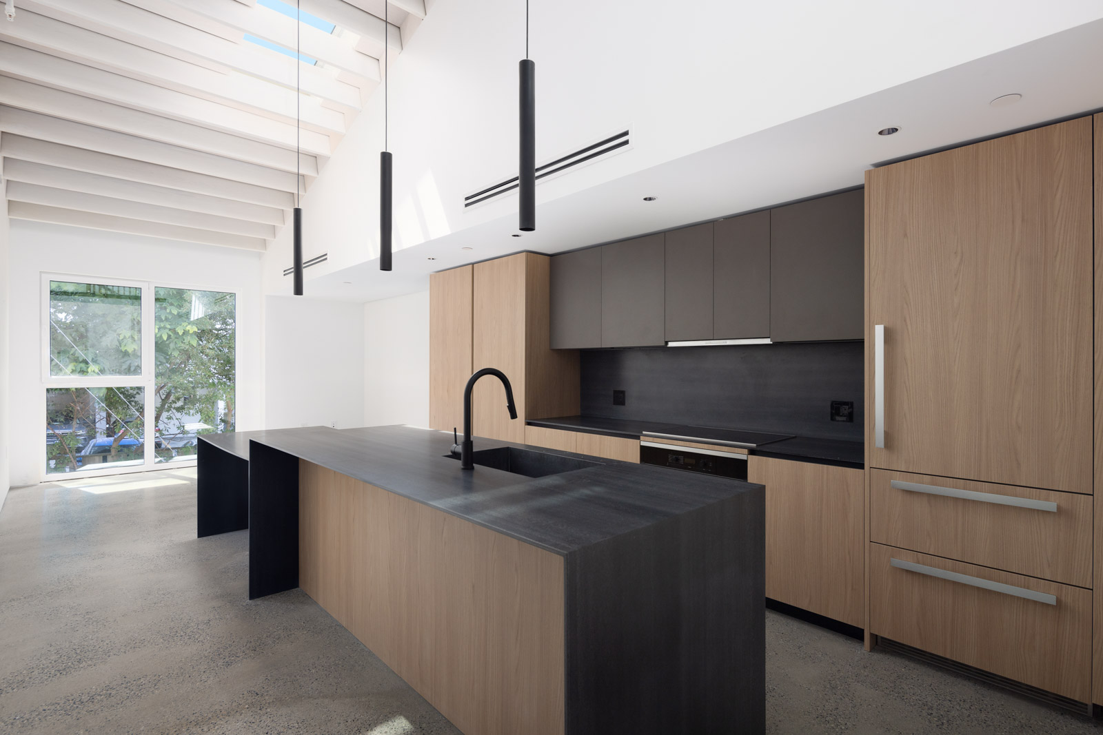 Modern kitchen with light wood cabinets, matte black countertops, built-in appliances, black fixtures, and large windows letting in natural light.