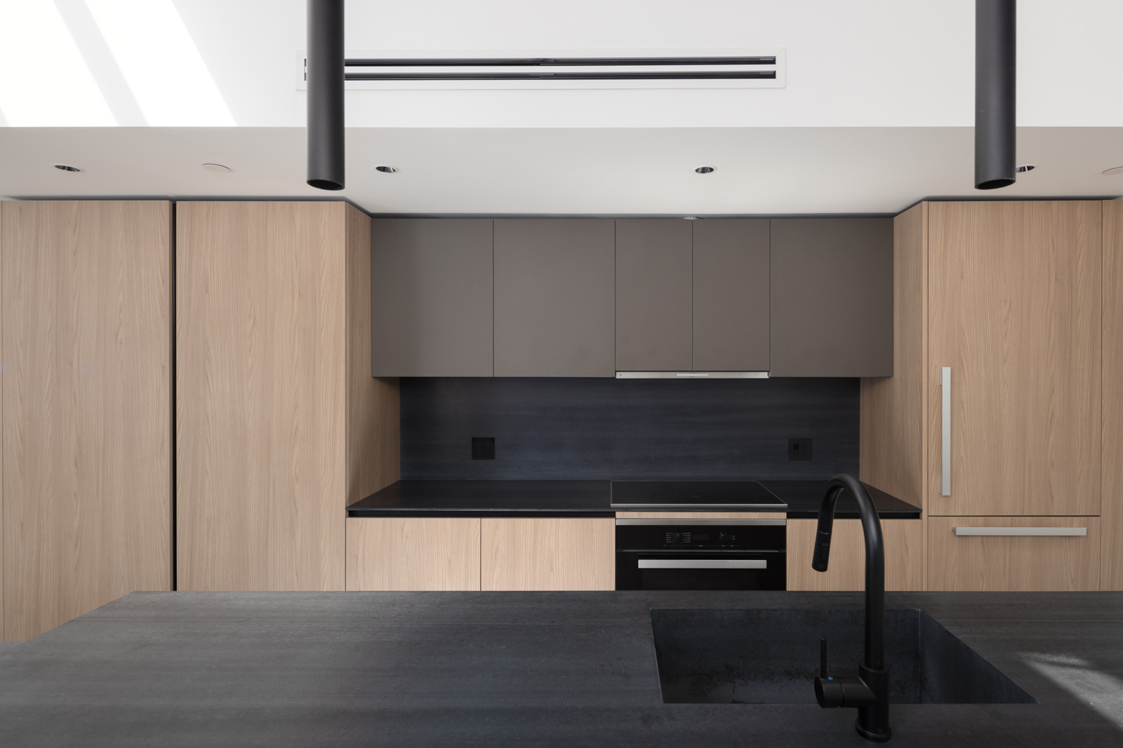 Modern kitchen with light wood cabinets, matte black countertops, built-in appliances, black sink, and minimalist fixtures under a bright skylight.