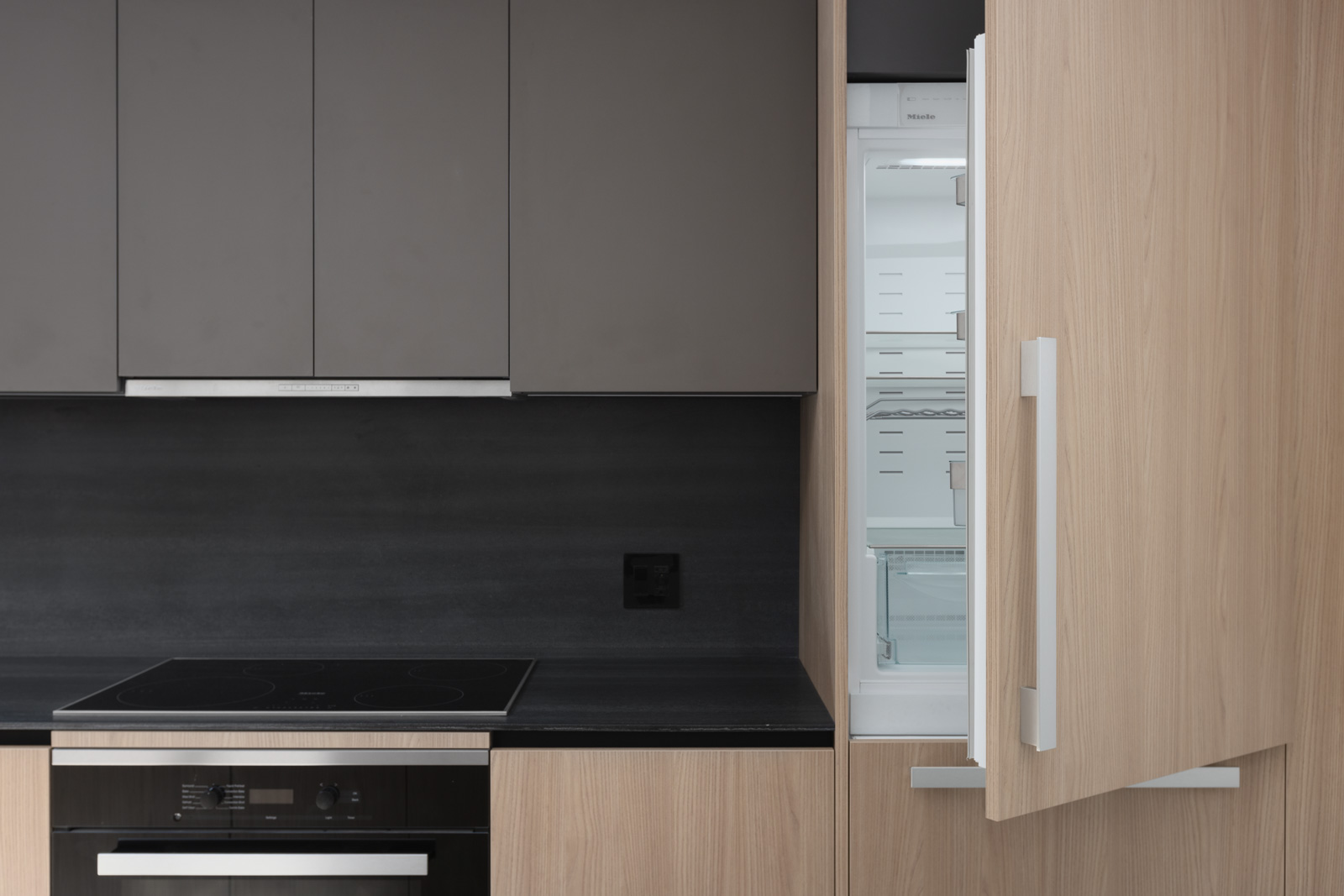Modern kitchen with dark cabinets, a black countertop, built-in oven, electric stovetop, and a light wood panel fridge with the door open.