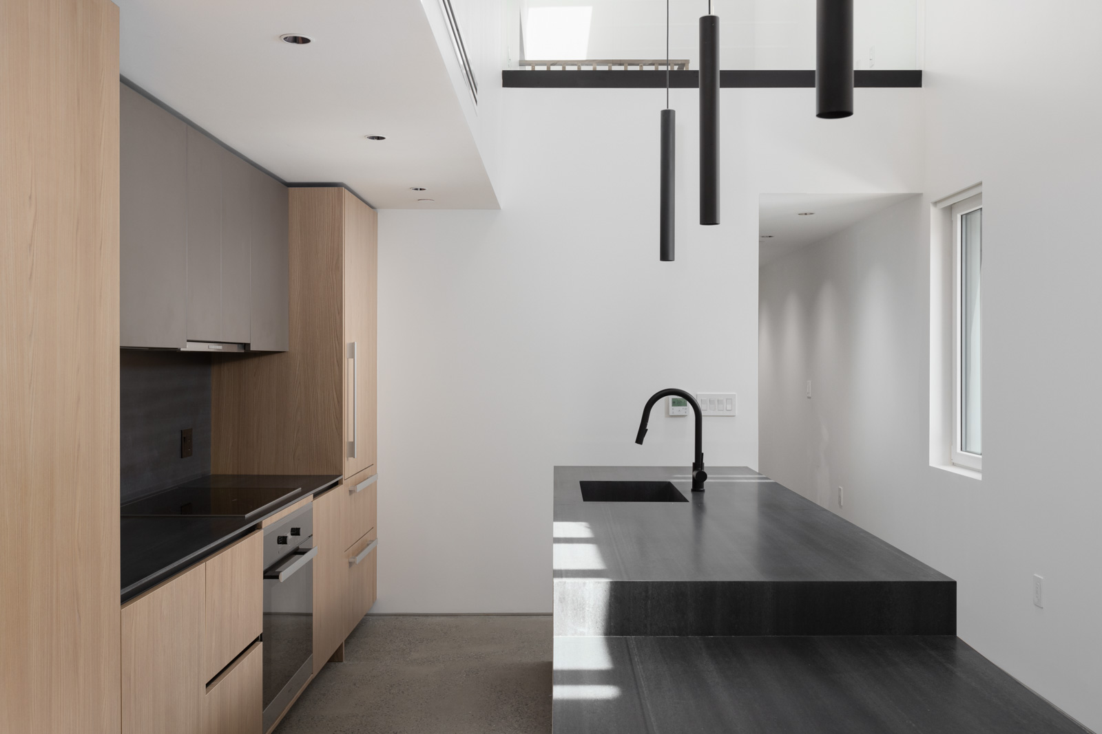 Minimalist kitchen with light wood cabinets, black countertop and sink, built-in appliances, and modern black pendant lights. White walls and natural light from a small window.