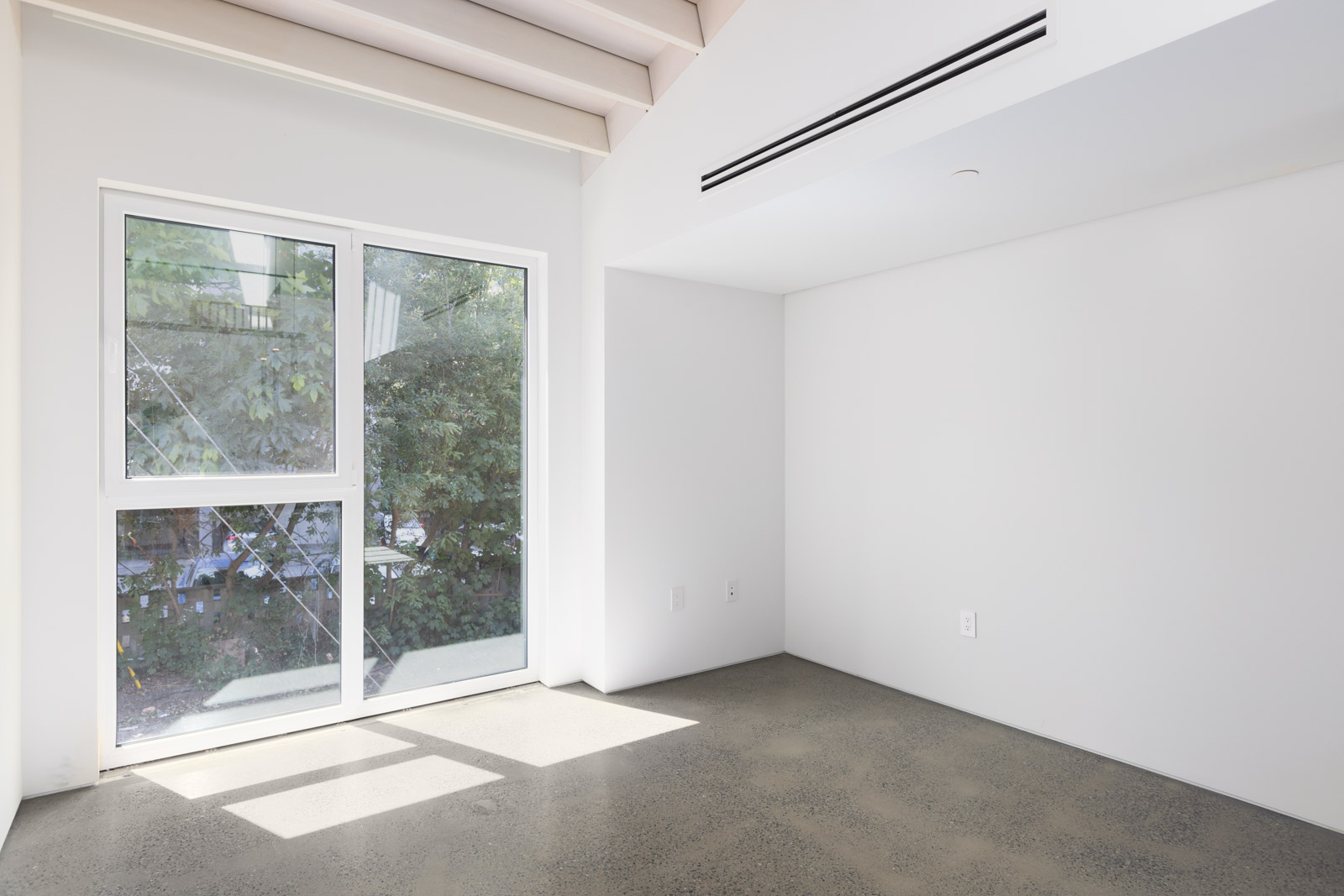 Empty room with white walls, a large window letting in natural light, polished concrete floor, and a ceiling air vent. Trees are visible outside the window.