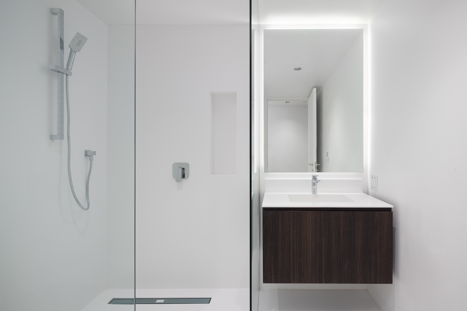 Minimalist bathroom with a glass-enclosed shower, wall-mounted showerhead, built-in shower niche, and a floating vanity unit with a sink and illuminated mirror.