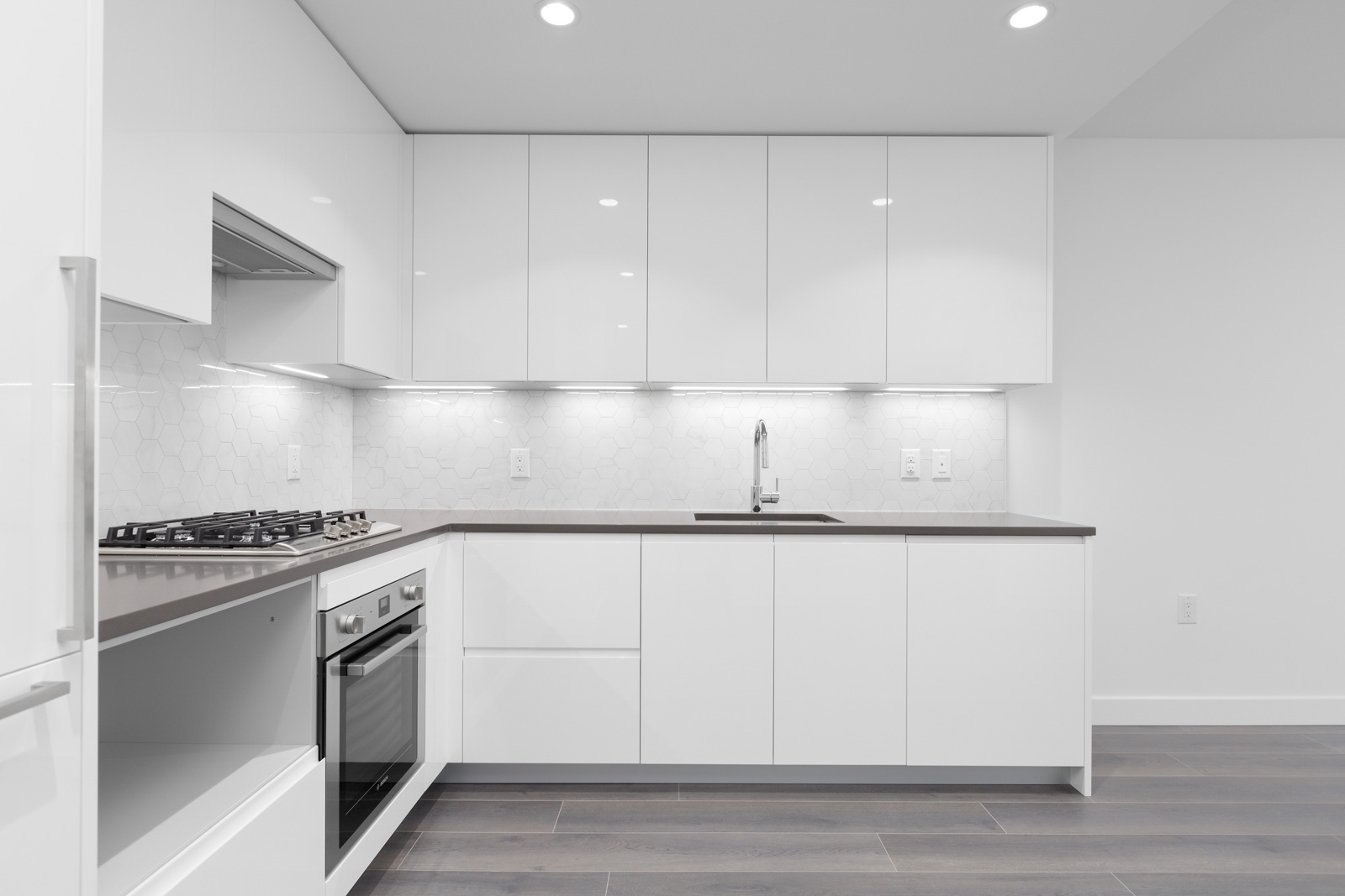 Modern white kitchen with built-in oven, gas stove, sleek cabinets, tile backsplash, under-cabinet lighting, and dark wood flooring.