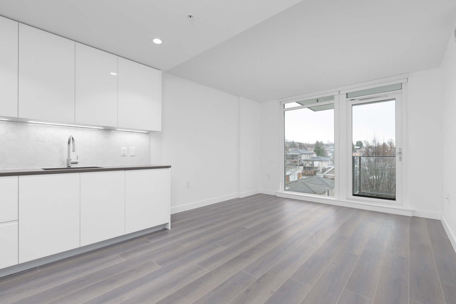 Modern, unfurnished apartment with white kitchen cabinets, grey wood flooring, and large windows leading to a balcony with a city view.