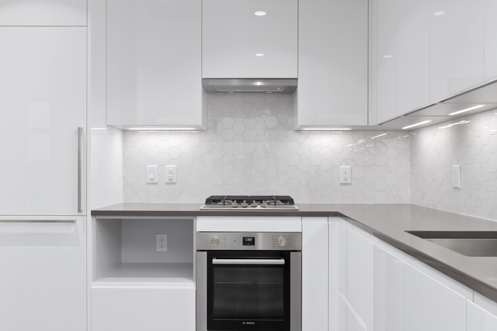 Modern white kitchen with glossy cabinets, hexagonal tile backsplash, built-in oven, gas cooktop, and under-cabinet lighting.