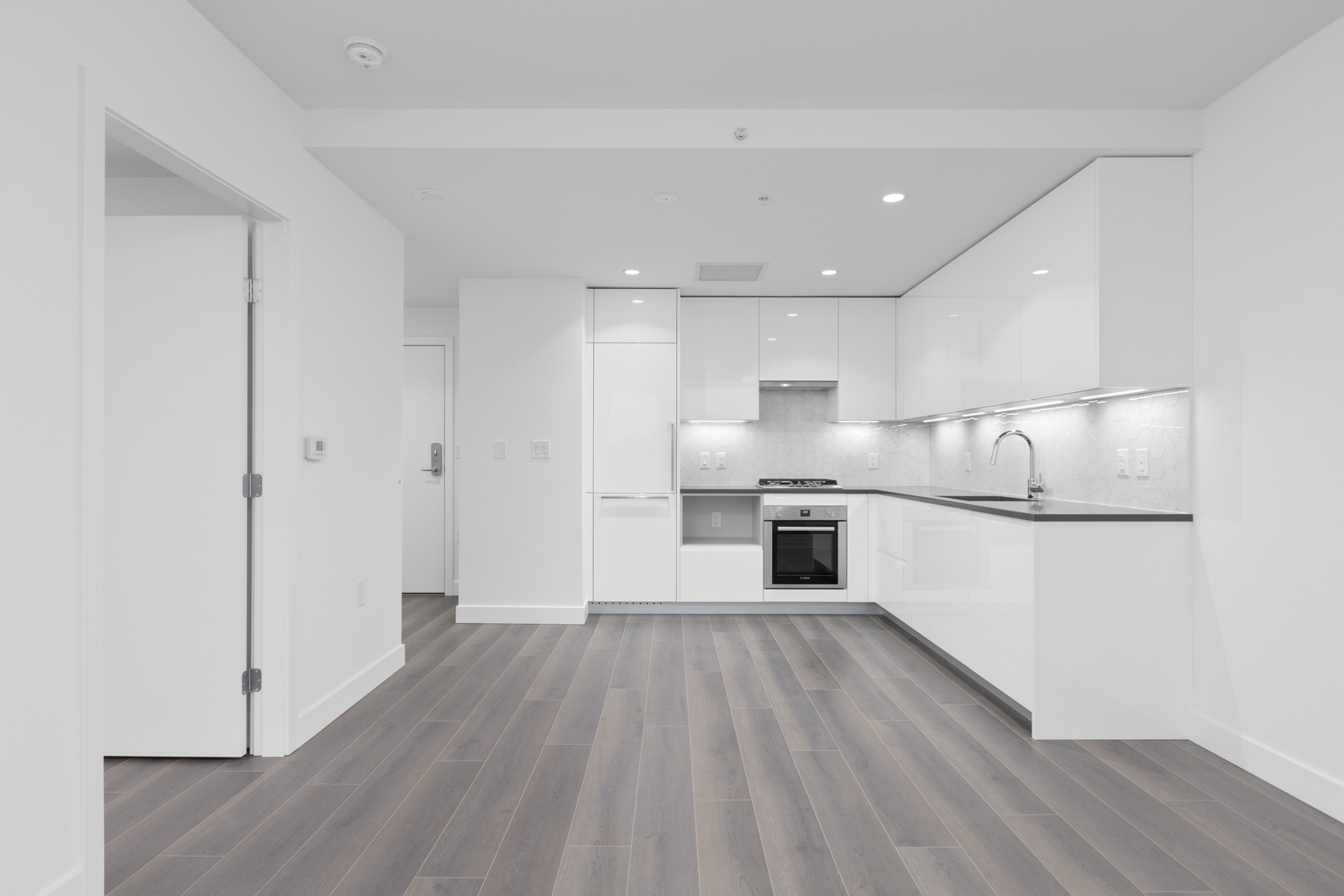 Modern, unfurnished kitchen with white cabinets, built-in oven, and light gray backsplash; open layout with wood-style flooring and white walls.