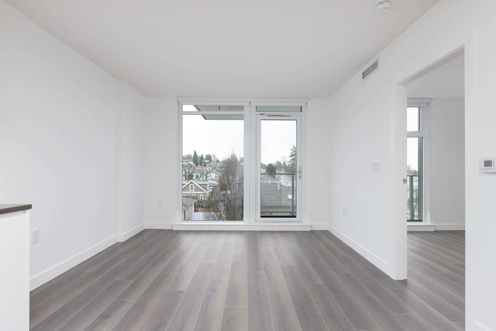 Empty, unfurnished apartment room with gray wood flooring, white walls, large windows, and a glass door leading to a balcony with a view of neighboring houses and trees.