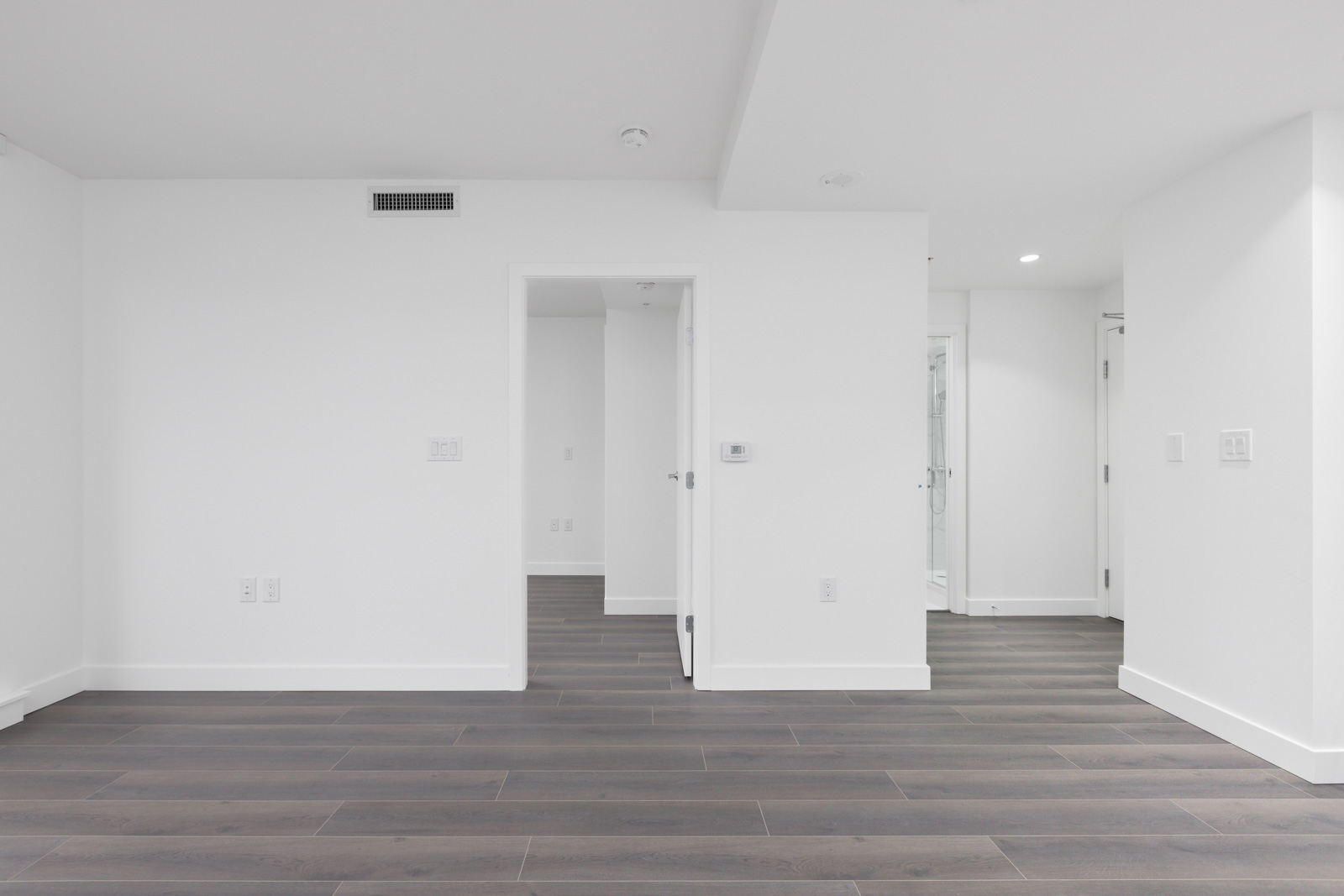 Empty modern room with white walls, dark wood flooring, and open doors leading to adjacent rooms; minimal lighting and no furnishings visible.