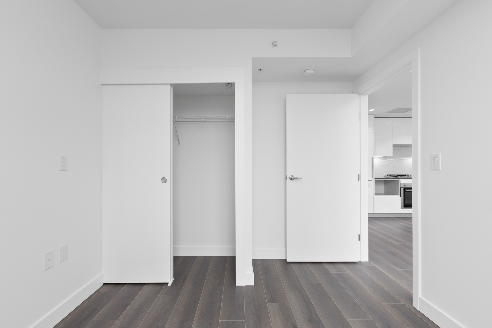 A minimalist, unfurnished room with dark wood flooring, a closet with sliding doors, an open door leading to a kitchen, and white walls.