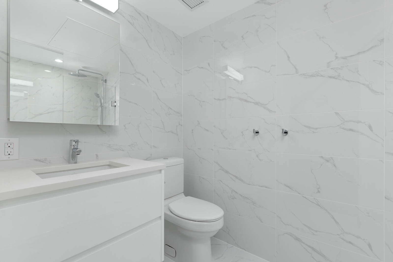 A modern bathroom with white marble-patterned tiles, a white vanity with sink, a wall mirror, and a toilet.